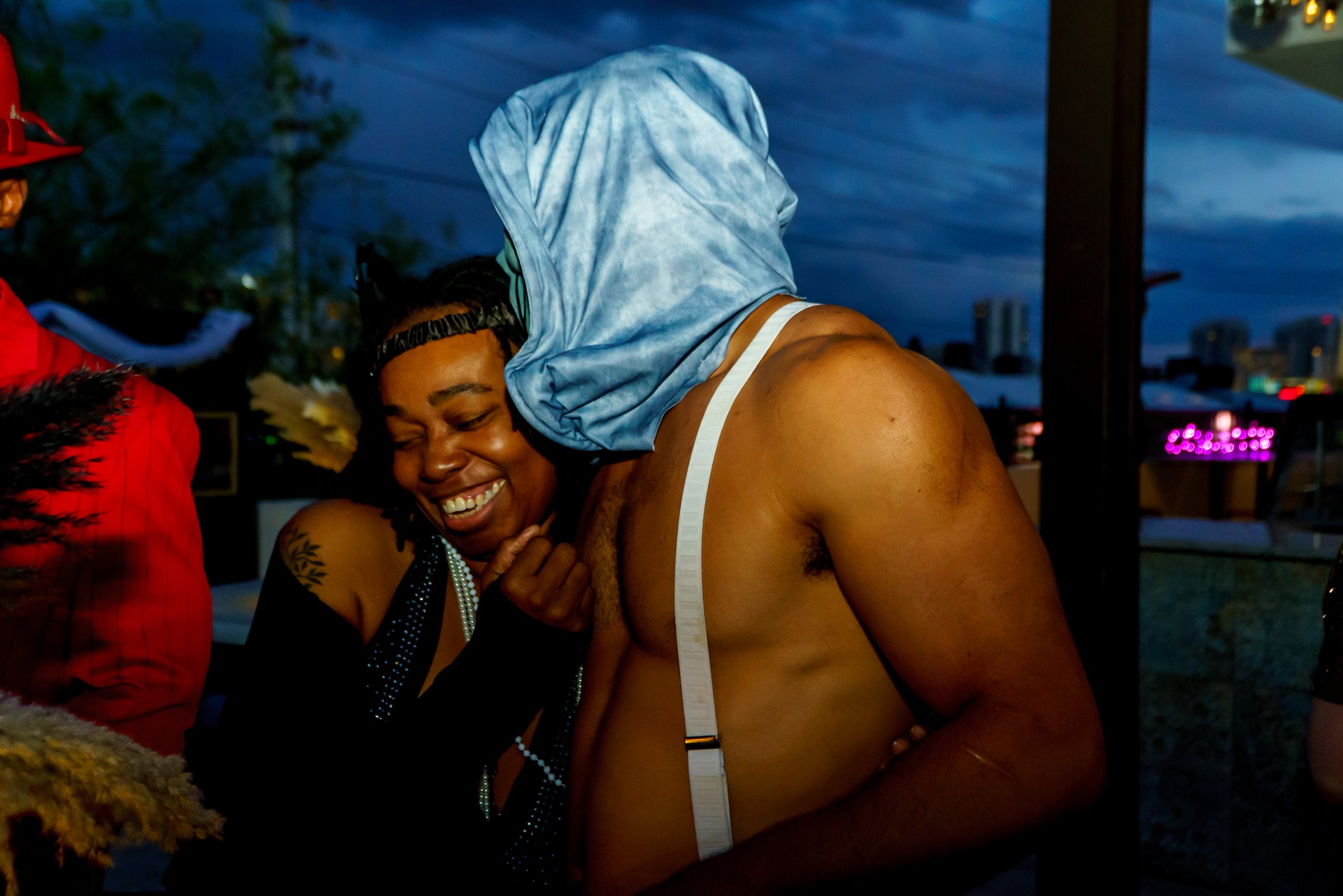 A woman smiling and wearing a blue bonnet and pearl necklace, leaning on a shirtless man with a measuring tape around his chest, outdoors during evening.