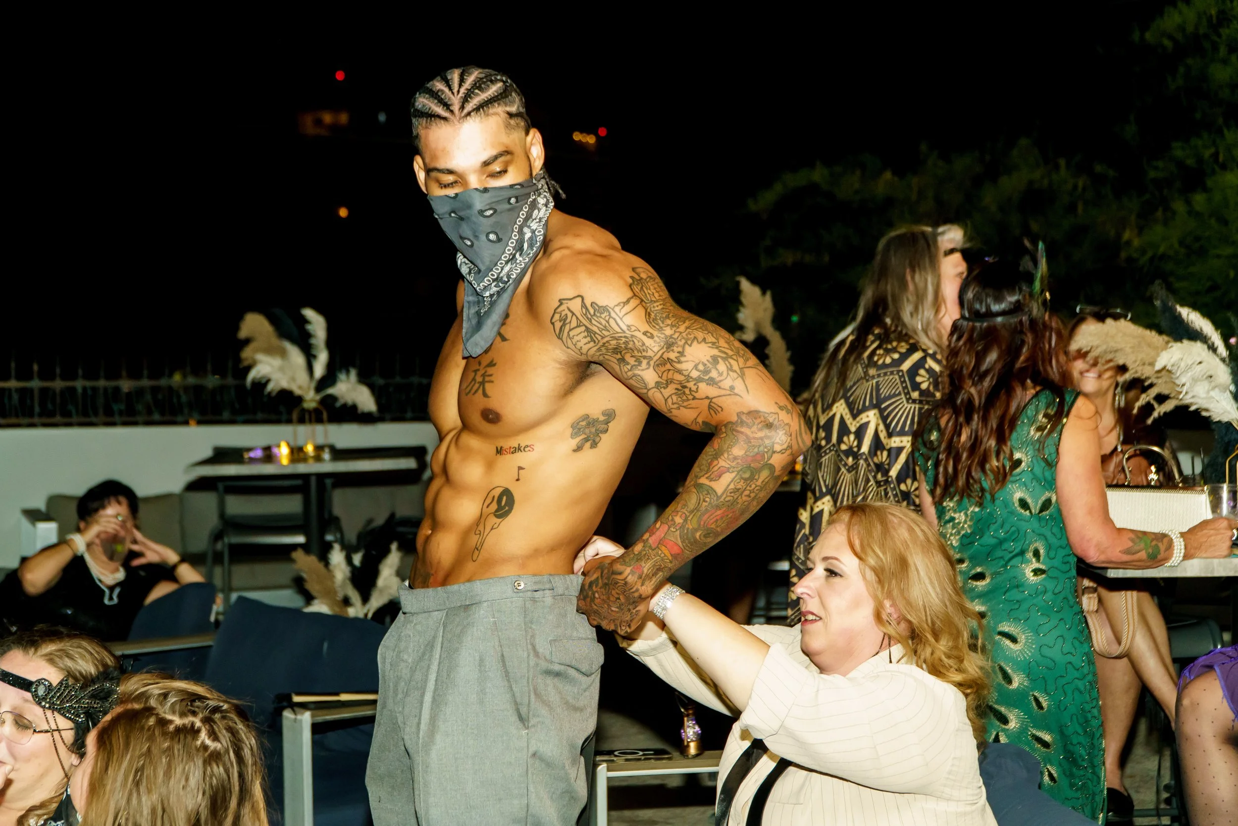 A shirtless man with tattoos and a bandana around his face dancing at a social gathering, surrounded by people sitting and talking.
