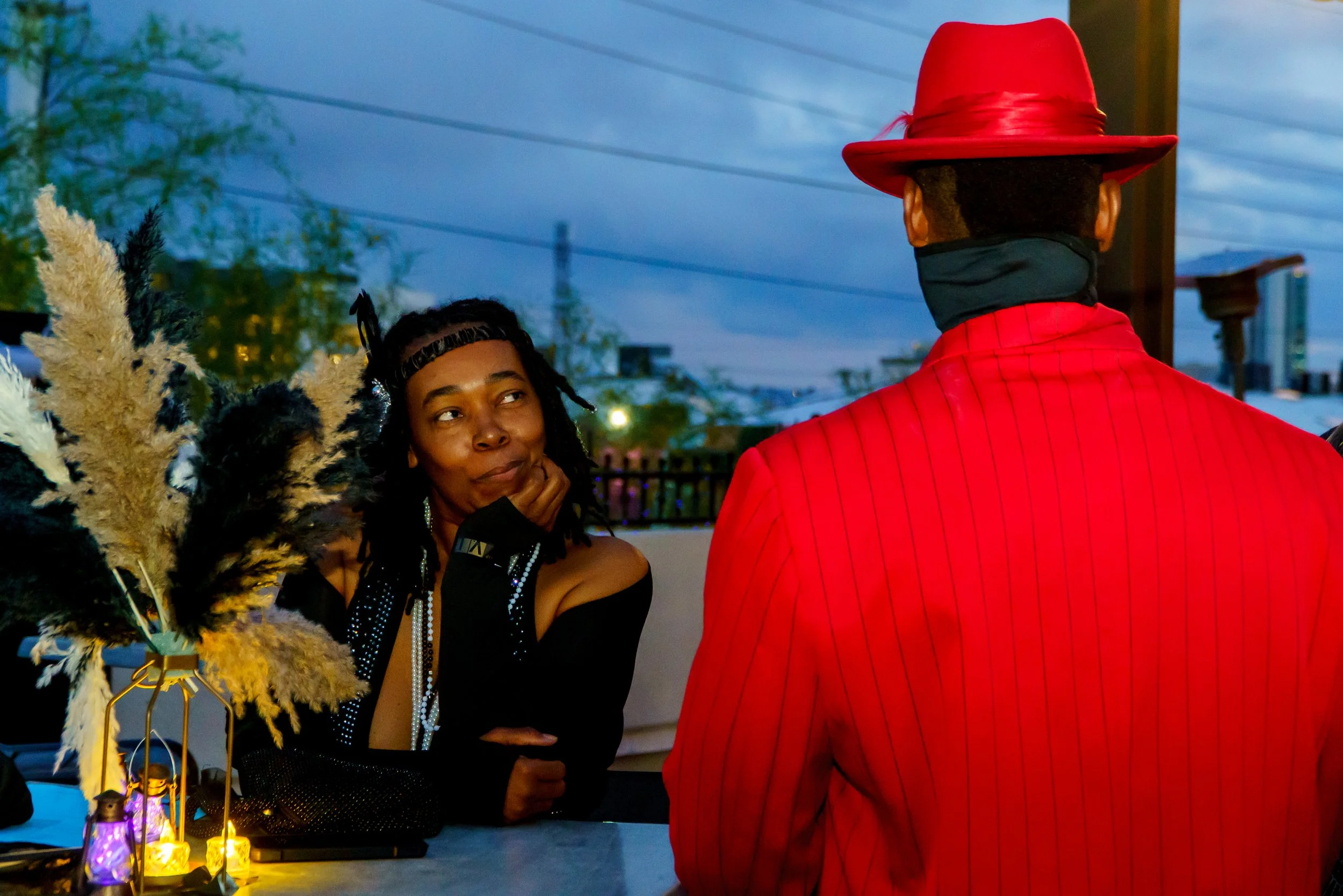 A woman with dreadlocks and a black off-shoulder top, wearing jewelry, observes a man dressed as a jazz musician in a red suit and red hat, during an evening outdoor event with decorative lanterns and string lights.