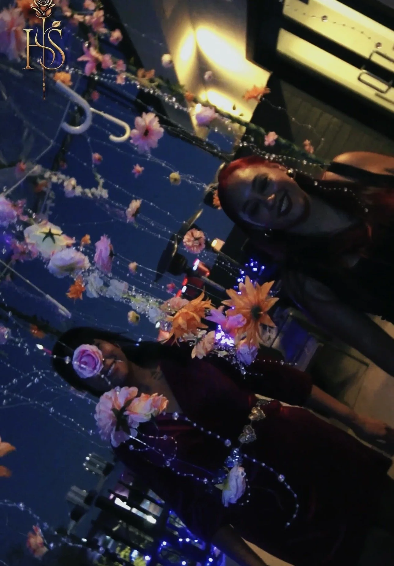 Nighttime street scene with two smiling women taking a selfie, surrounded by hanging flowers and decorative lights.