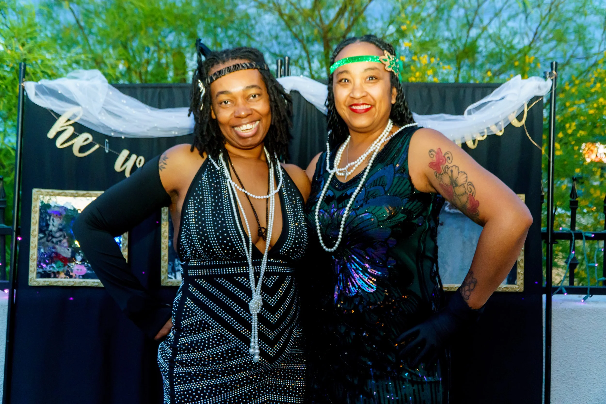 Two women smiling and posing outdoors in front of a dark backdrop with white tulle and gold lettering, surrounded by green trees, dressed in black party dresses with pearl necklaces and headbands.