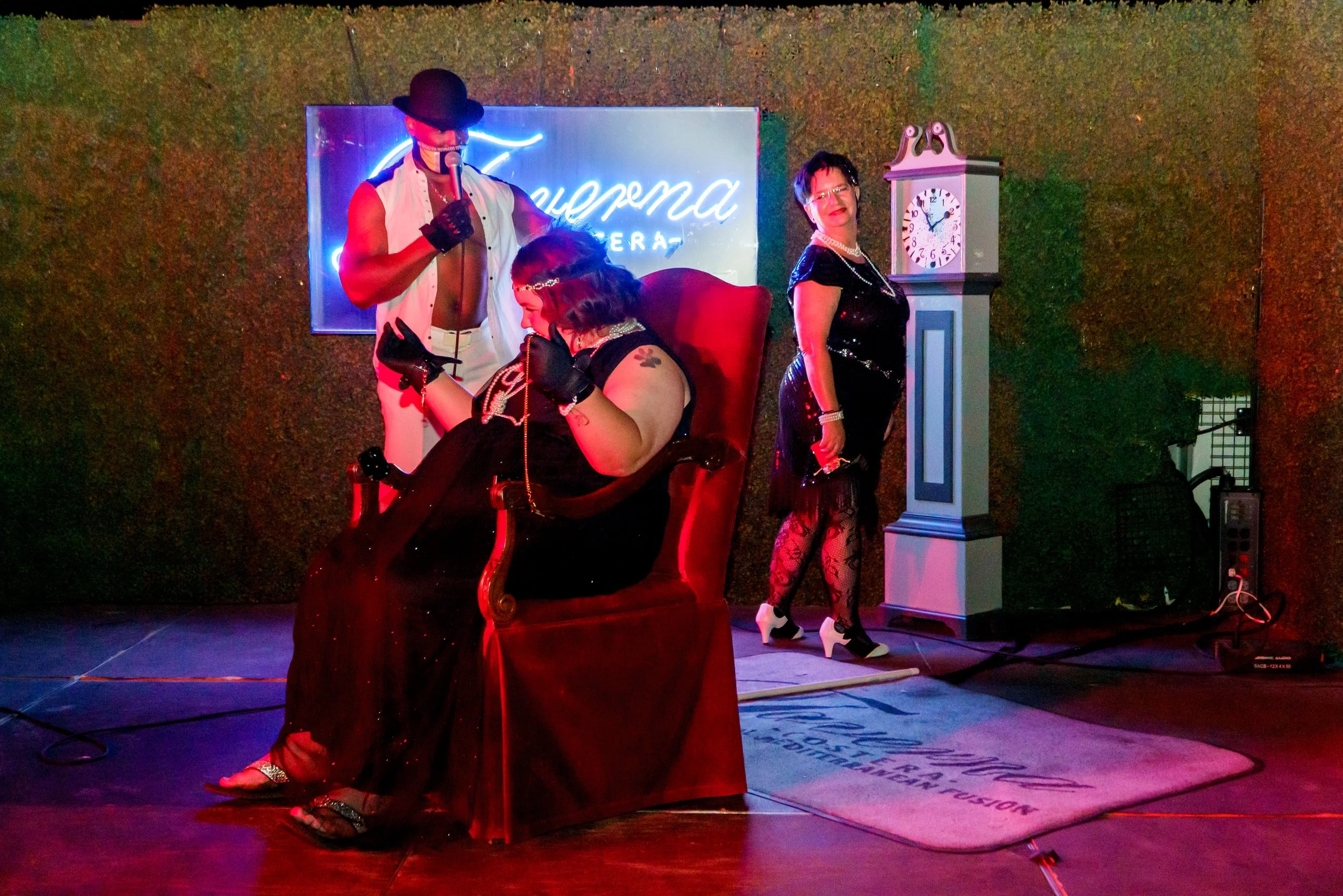 A person sitting in a red chair with a woman standing beside them, both dressed in vintage-style black outfits with pearls, on a stage with a neon sign in the background, a grandfather clock, and colorful lighting.
