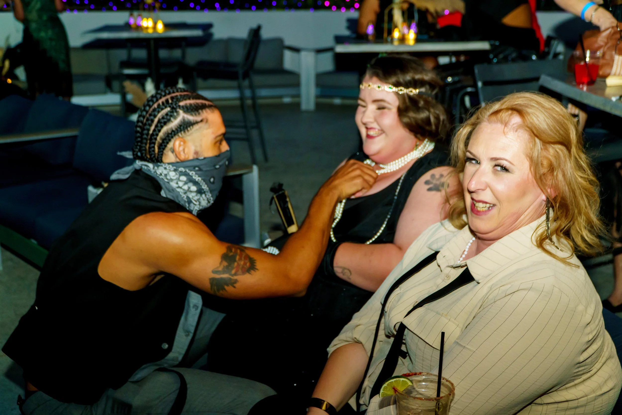 A group of three women at a social gathering, one with braided hair and tattoos, smiling and talking to two women with light skin, one with curly blonde hair and the other with dark hair with a headband, all engaged in conversation in a dimly lit ind