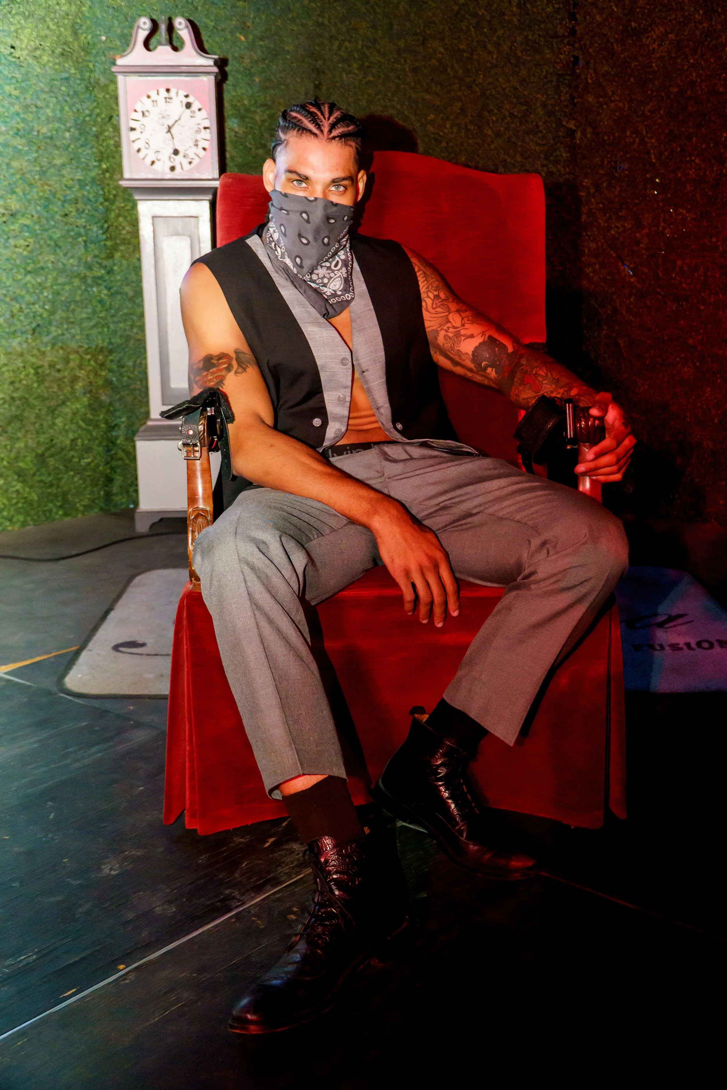 Person sitting in a red chair with tattoos on their arms, wearing a face covering, gray pinstripe pants, and black boots. There is a clock on the wall behind them and a green textured backdrop.