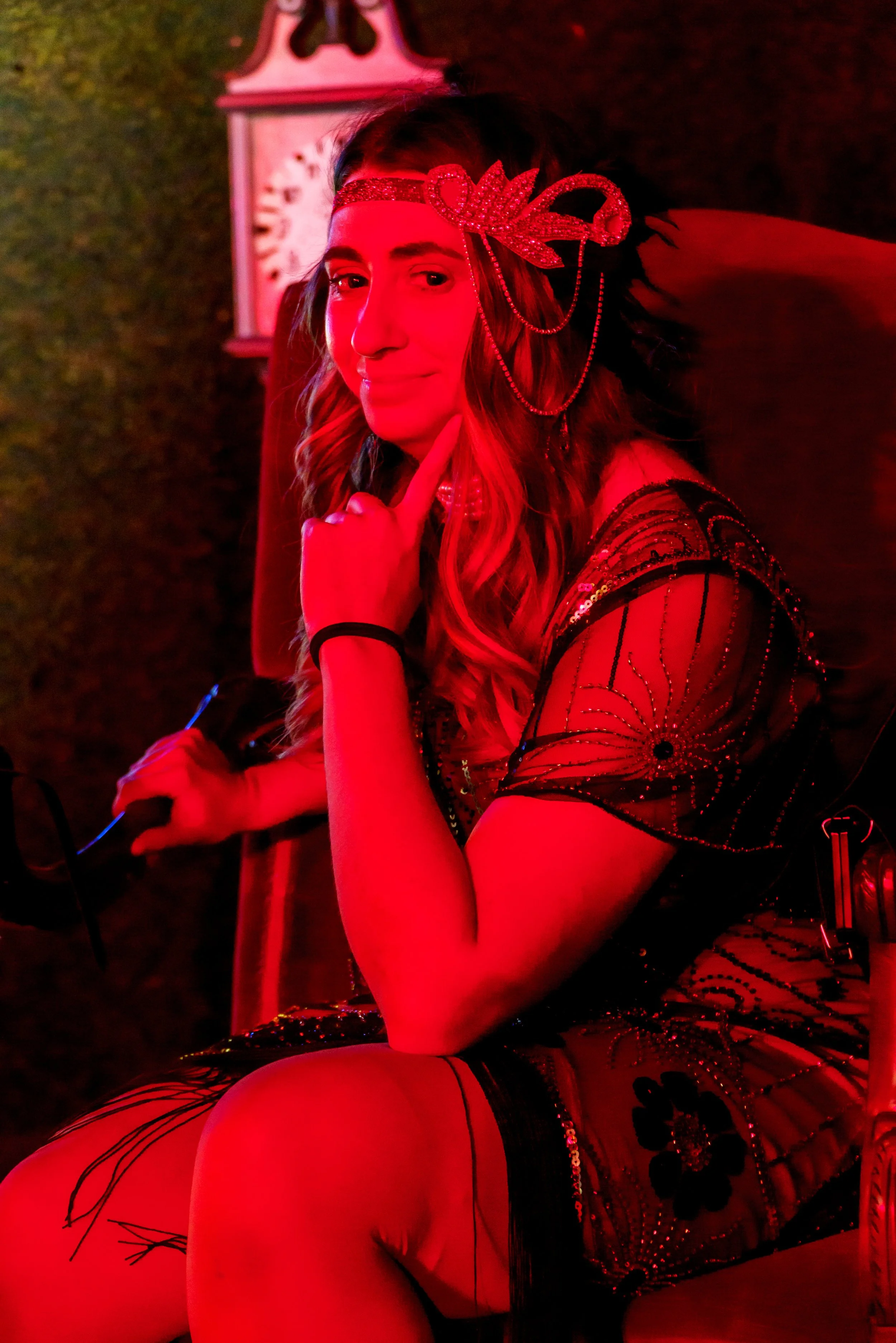 A woman with long, wavy hair, wearing a decorative red headband with beads and feathers, sitting in a dimly lit environment illuminated by red lighting. She is dressed in a black, embellished outfit with long sleeves, and her pose suggests she is pos