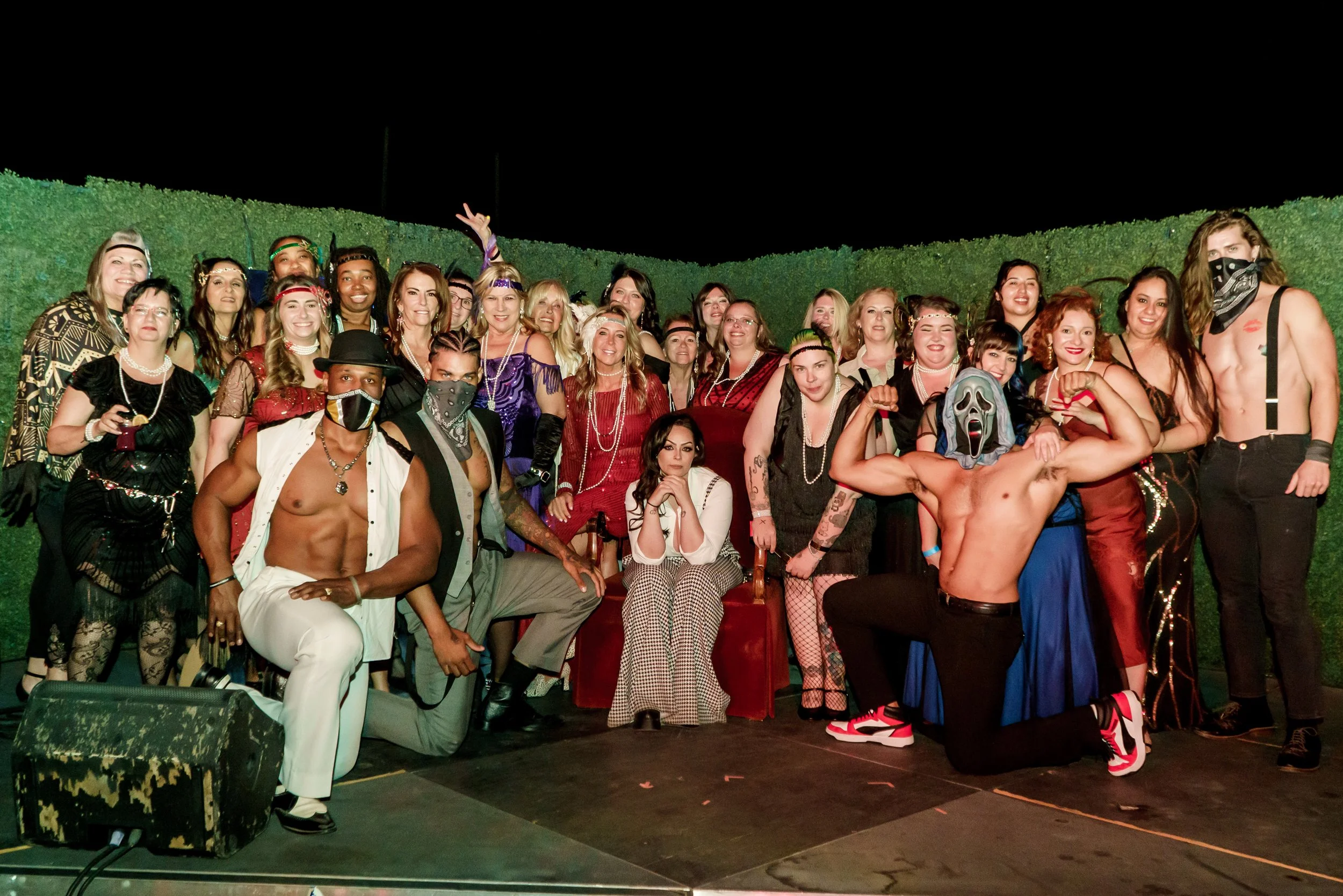 A large group of people dressed in various costumes, some with masks and accessories, gathering on a stage with a green hedge background and a dark night sky.