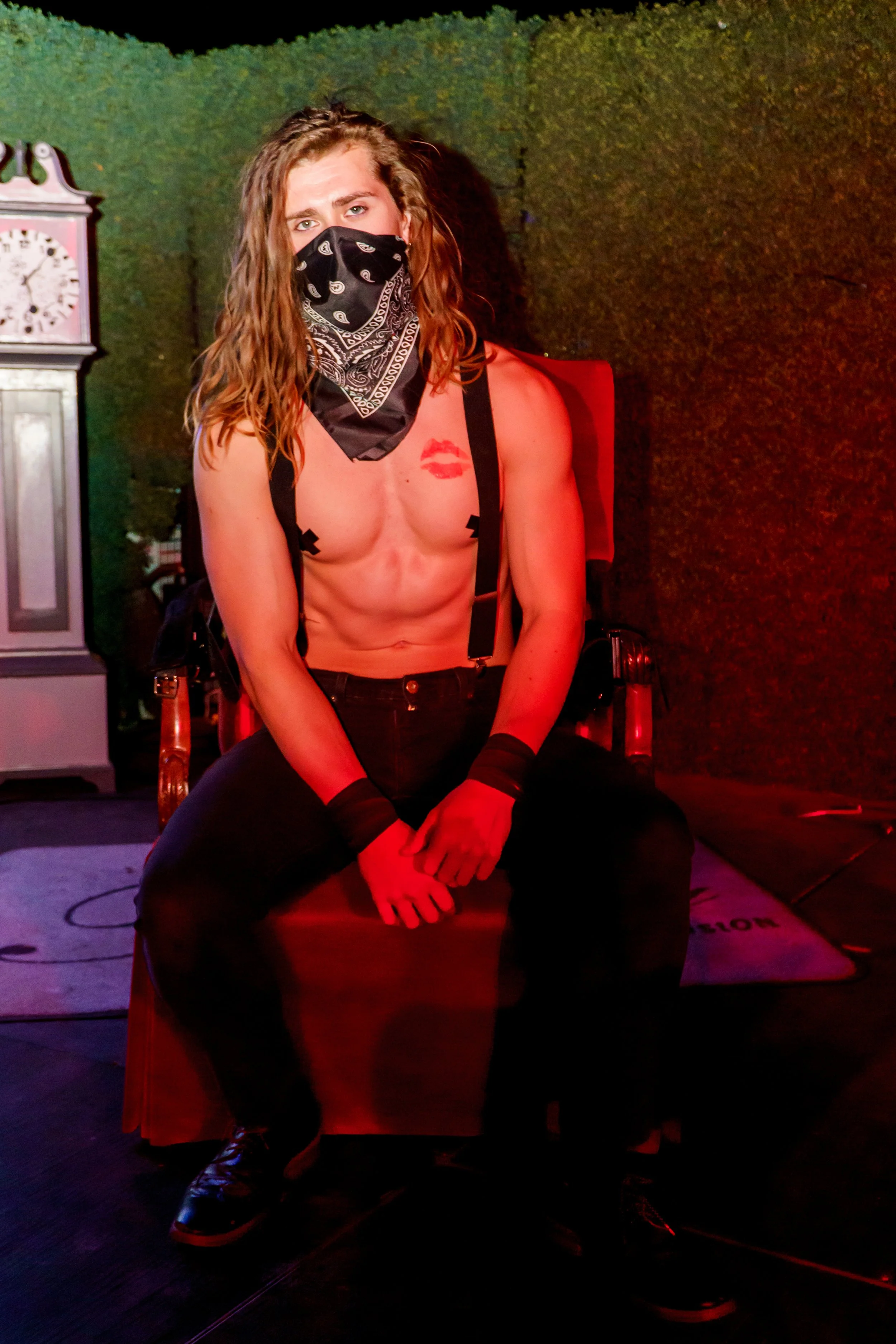 A person with long curly hair wearing a bandana mask covers mouth and nose, black suspenders, black pants, and black shoes, sitting with legs apart on a wooden chair in front of a colored background, with a painted lips tattoo on chest and black tape