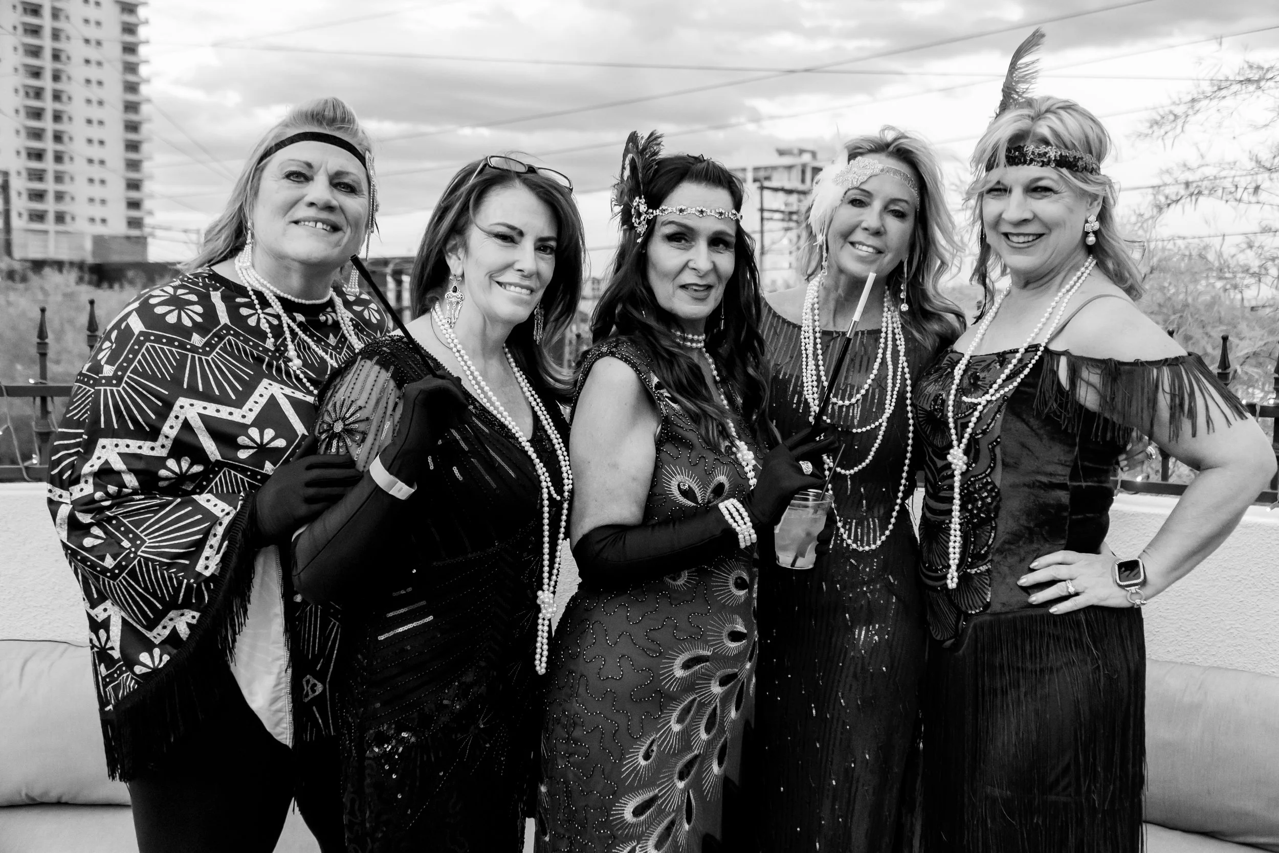 A group of five women dressed in vintage 1920s flapper costumes, with headbands, feather accessories, and pearl necklaces, standing outdoors with city buildings and power lines in the background.
