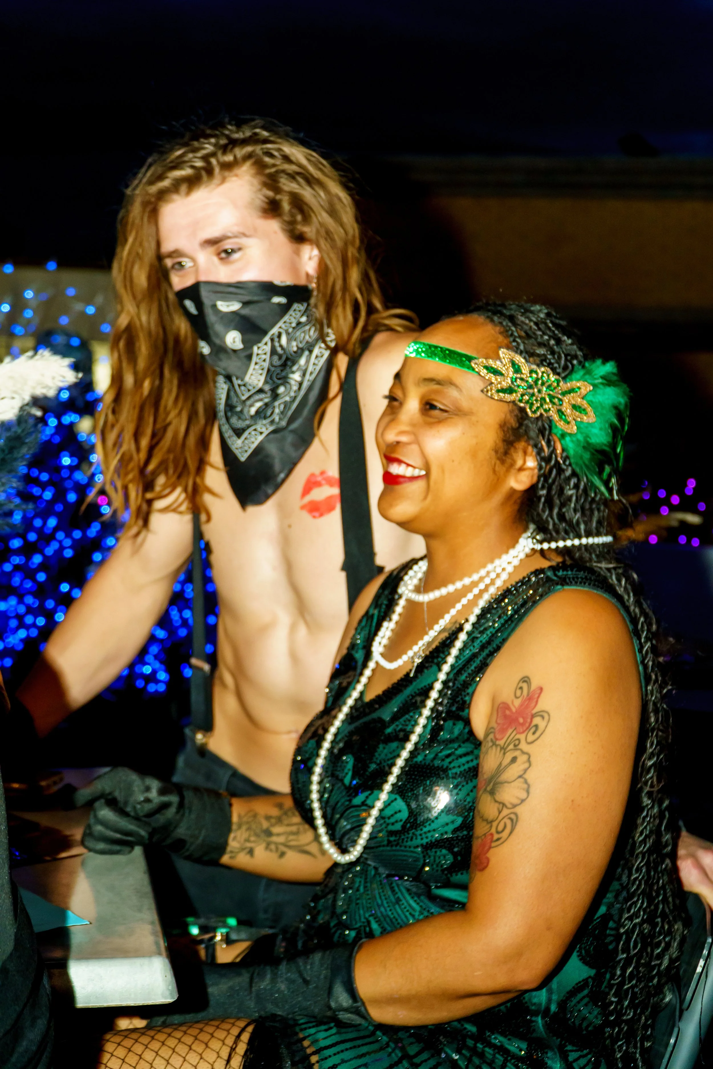 Two women, one with long curly hair and a face mask, the other with braided hair and a green sequined headband, smiling at a party with Christmas lights in the background.