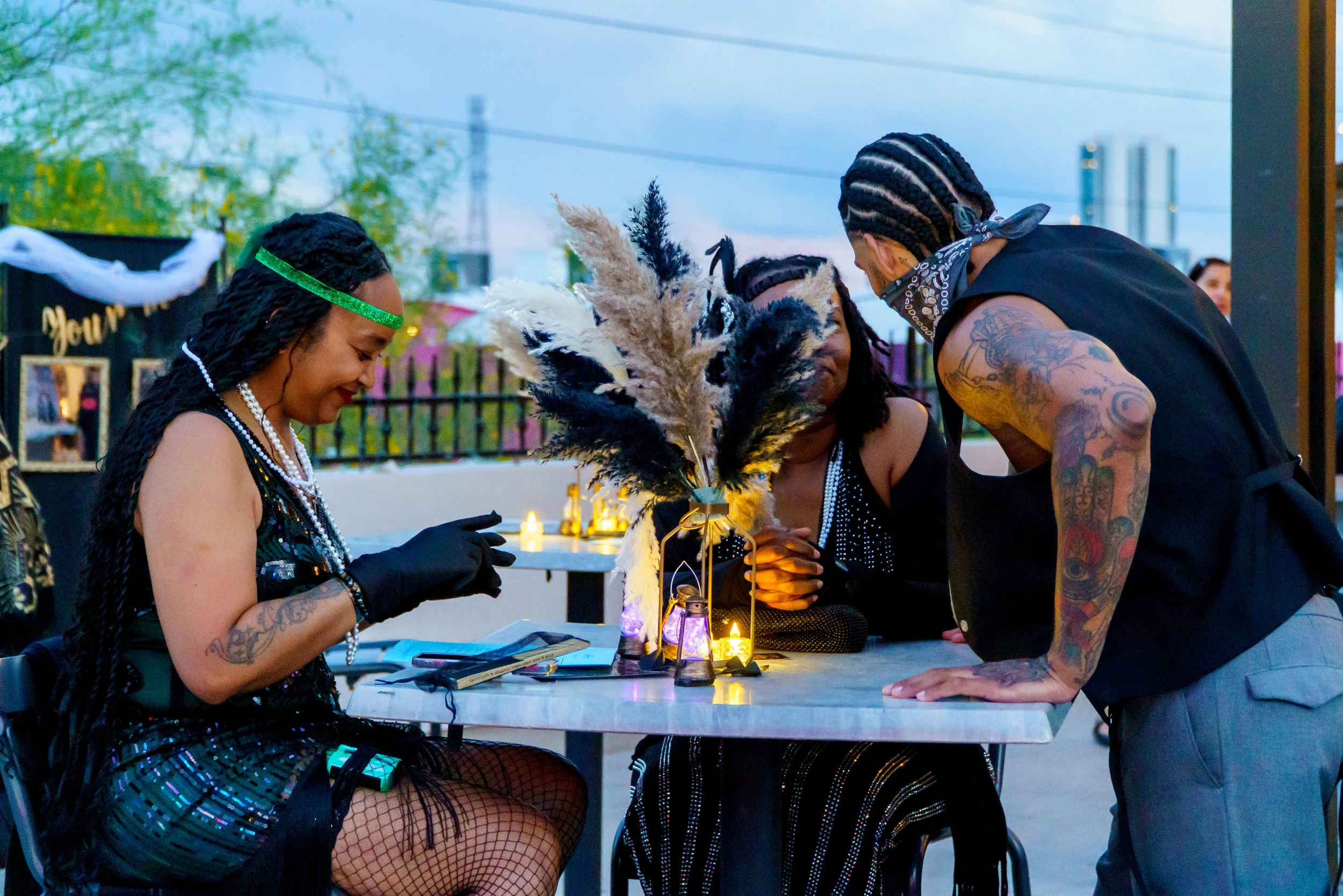 Group of people at a social gathering outdoors around a table with candles and decorative feathers, engaging in conversation and looking at items on the table.