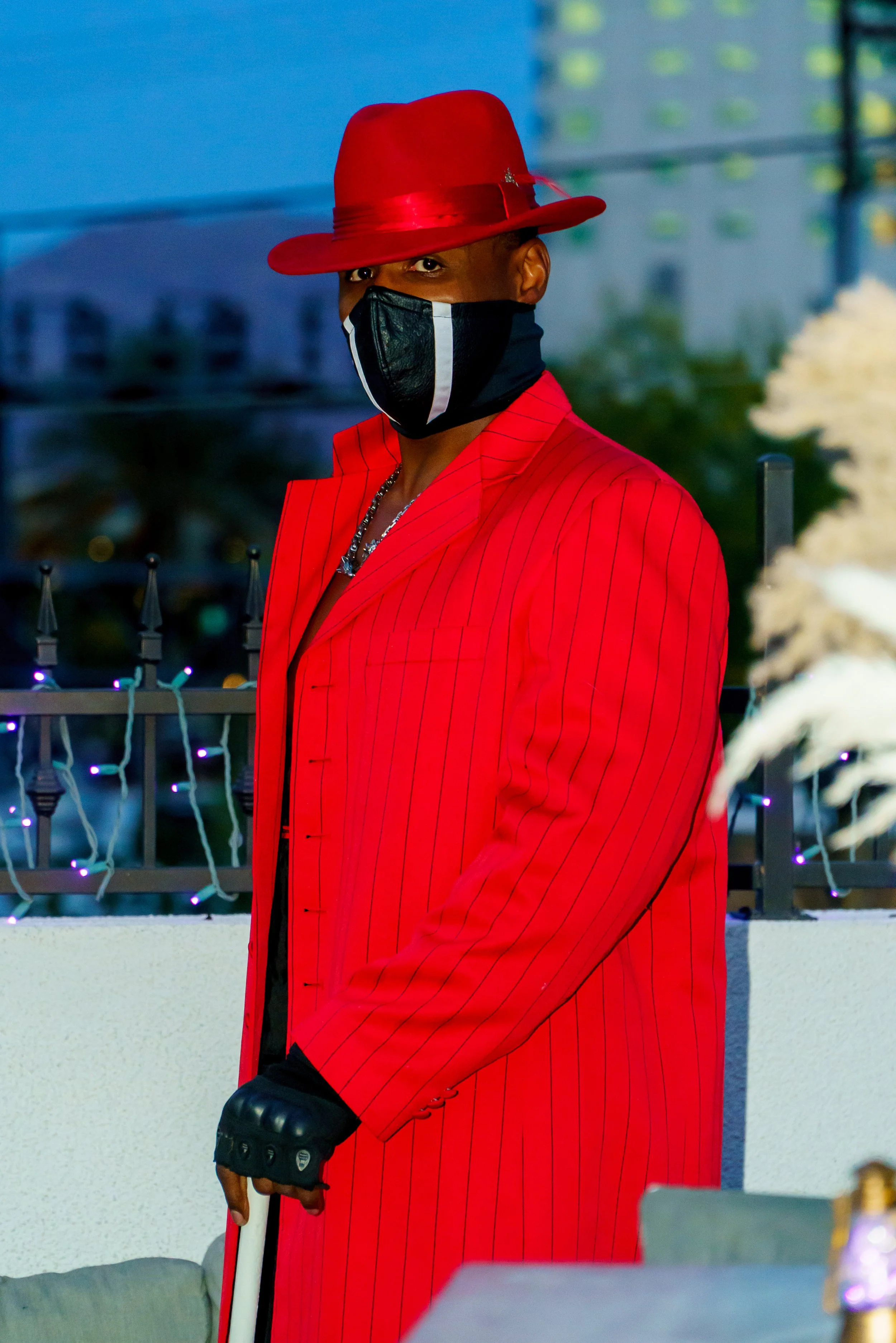 Person wearing a red pinstripe suit and a matching red hat, with a black face mask, standing outdoors during evening, holding crutches, with city buildings and decorative string lights in the background.