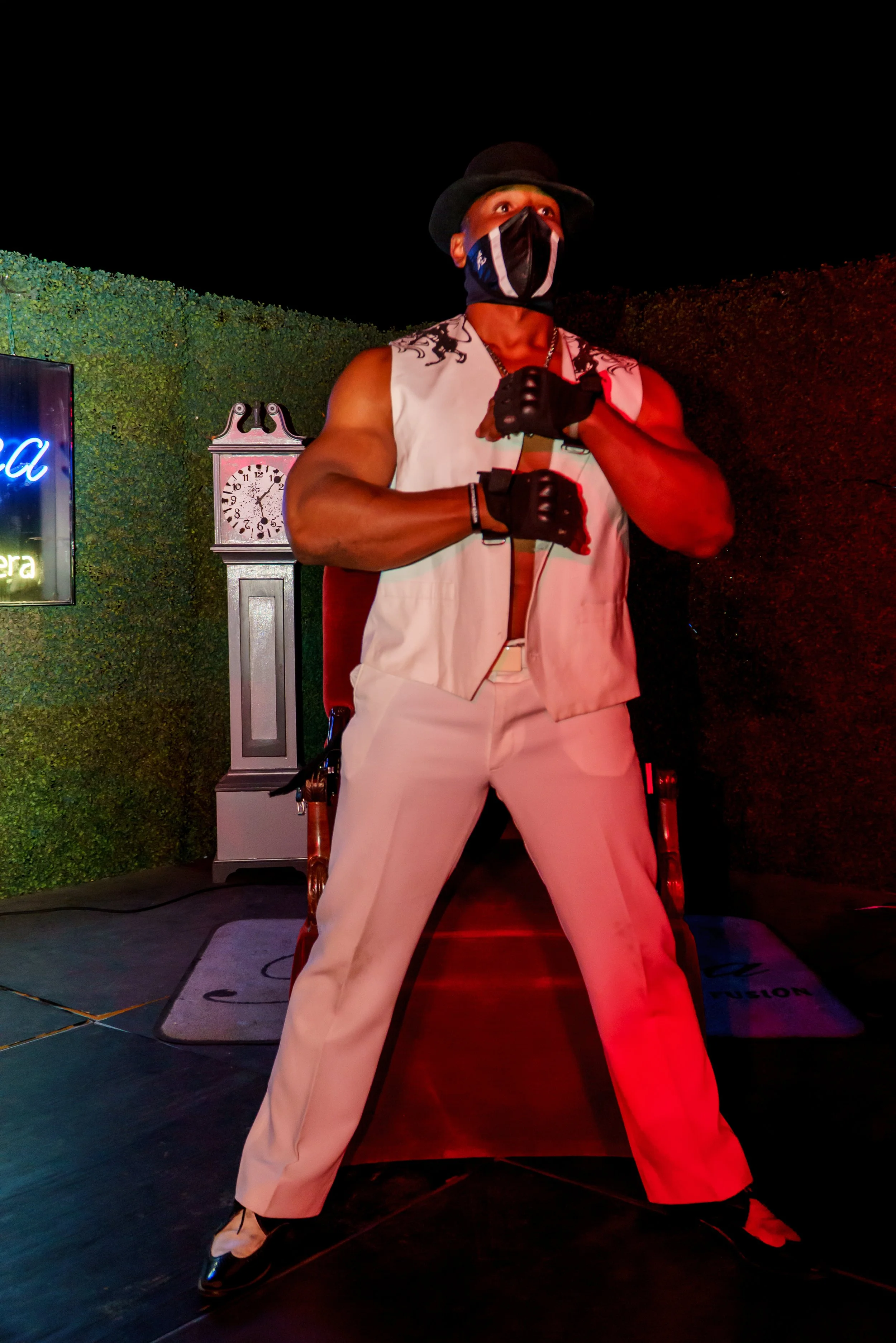 A man in a white sleeveless vest, white trousers, and black shoes, wearing a black hat, black gloves, and a black face mask, stands with legs apart and fists raised in a boxing pose on a stage with a clock in the background.