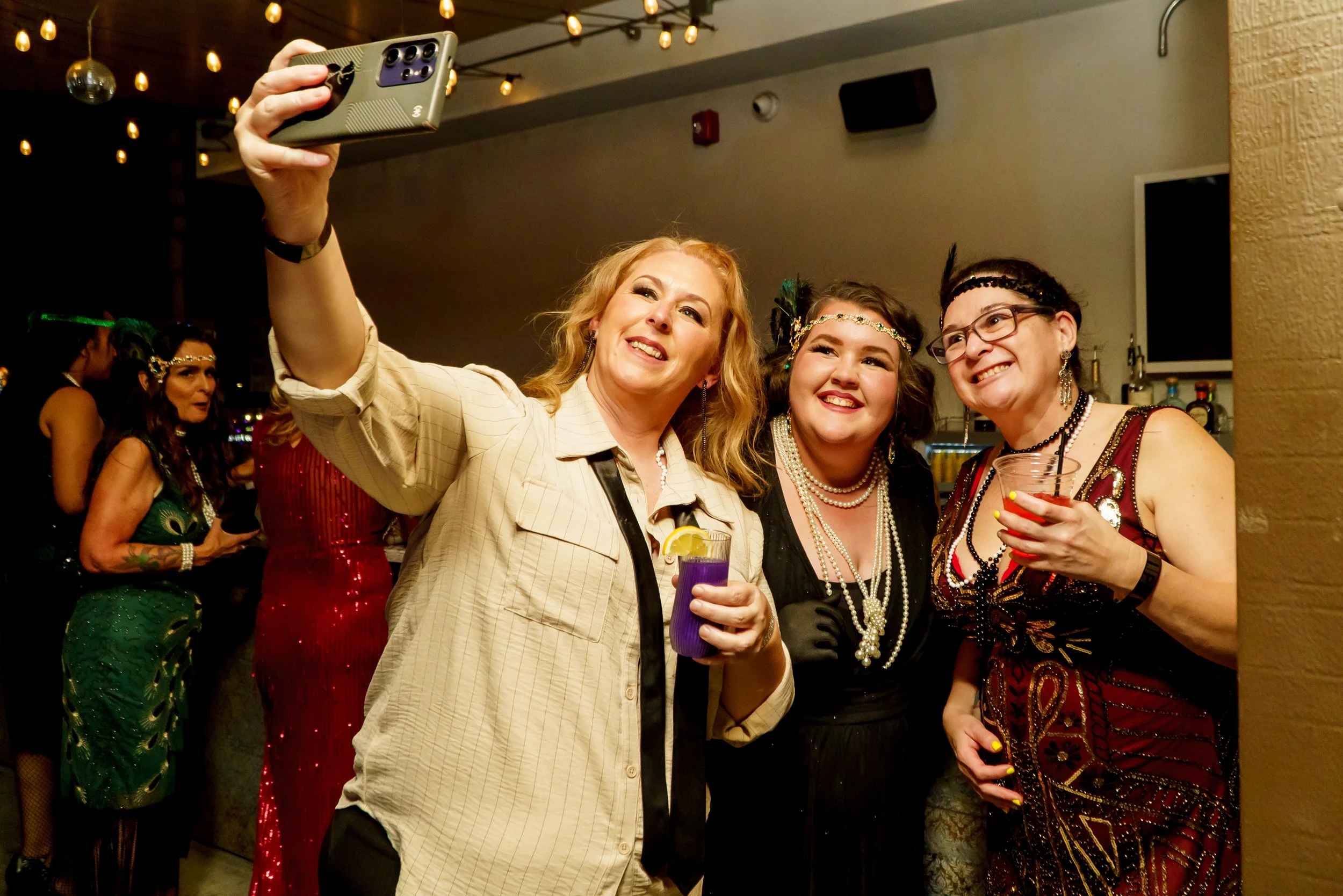 Group of women dressed in 1920s style flapper costumes taking a selfie together at a party, with some women in the background also dressed in costumes and dancing.