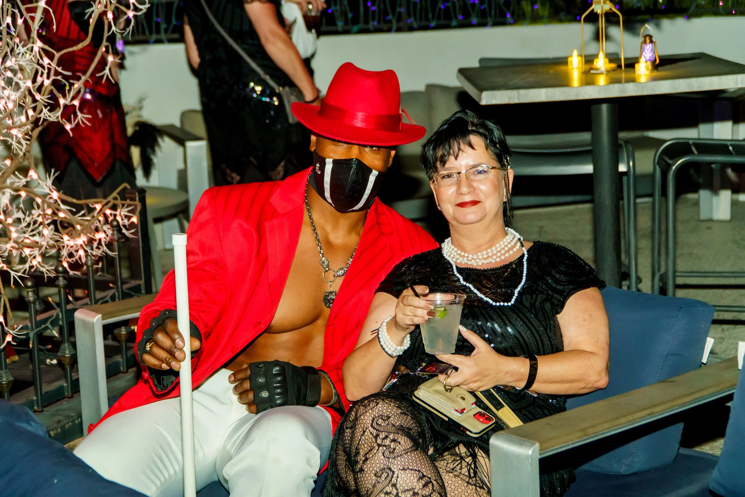 Two women sitting together at a social gathering, one wearing a red hat and mask, the other in a black dress and glasses, holding a drink.