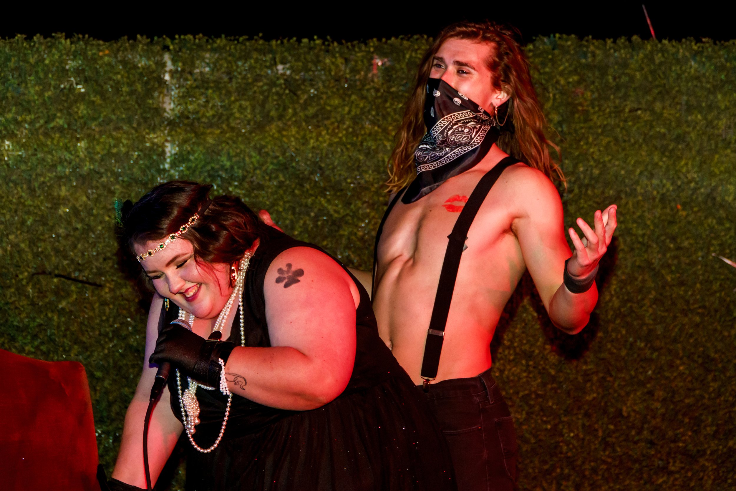 Two women performing at night, one with a mask and lipstick kiss mark, wearing suspenders and making a hand gesture, while the other is singing into a microphone, against a background of greenery.