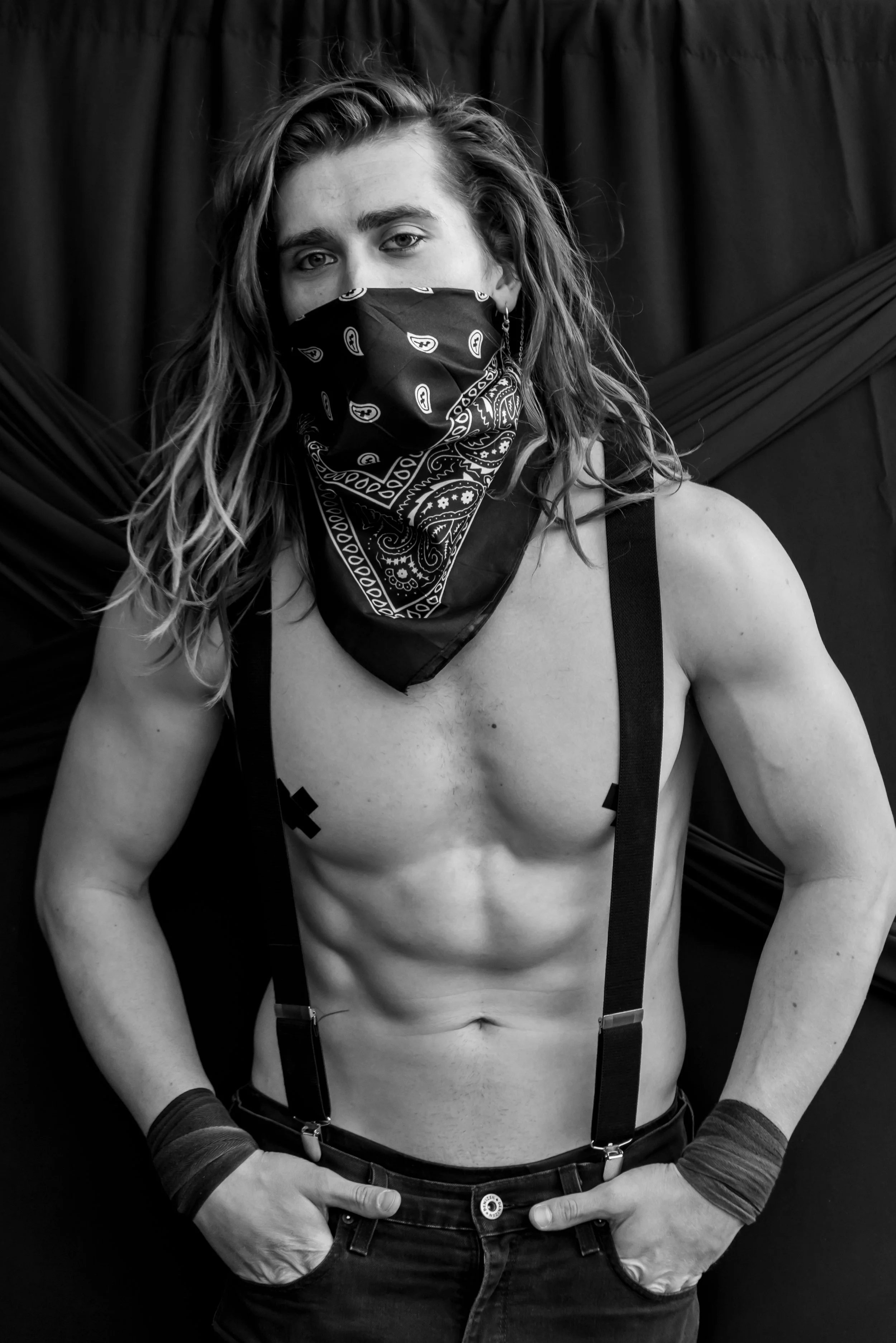 Black and white photograph of a shirtless man with long hair, wearing a bandana face mask, suspenders, and gloves, standing in front of a dark curtain.