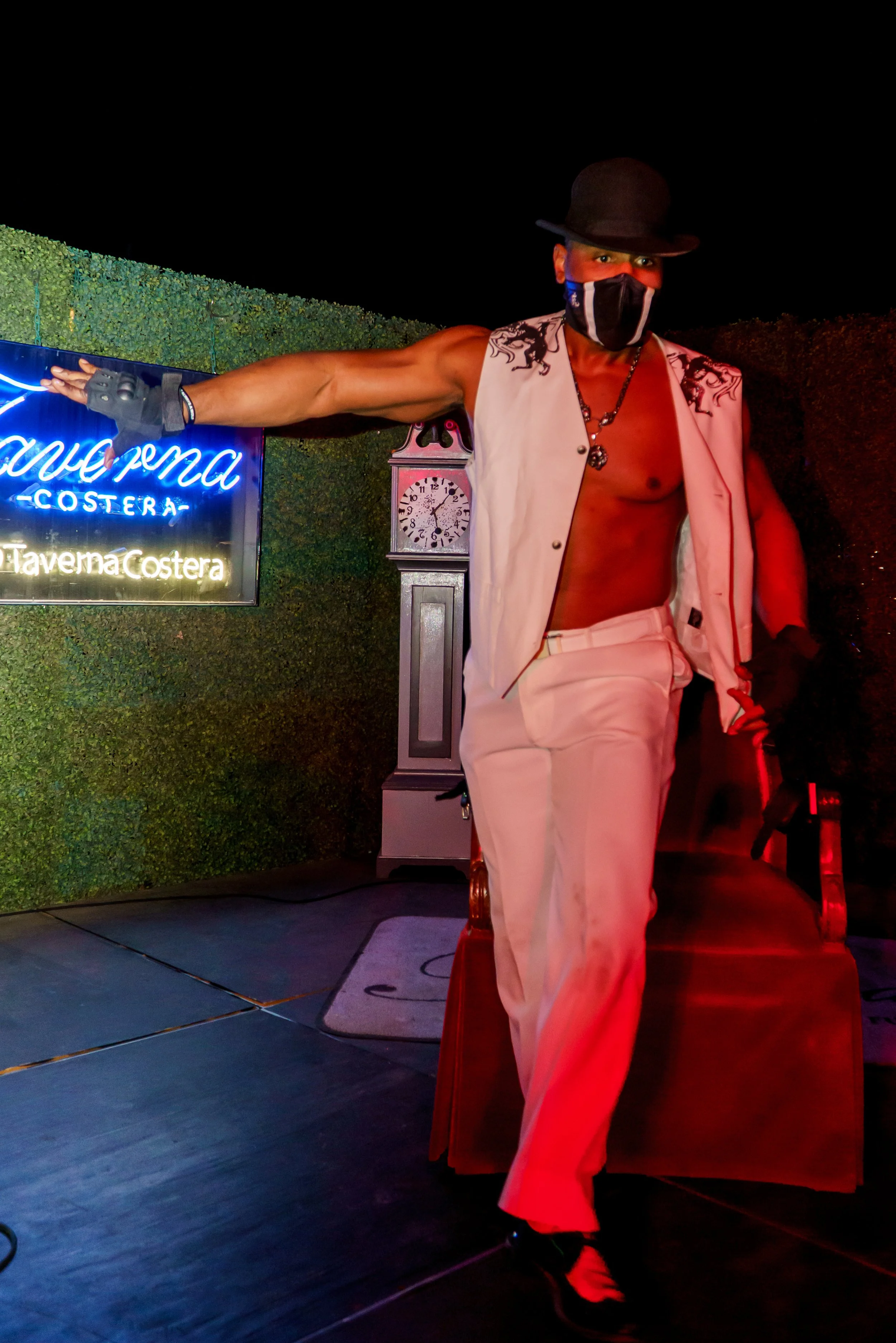 A shirtless man with long curly hair wearing a black hat, a face mask, a sleeveless jacket, white pants, and a chain necklace, standing in front of a green hedge at night near a neon sign that reads 'Taverna Costera' and a vintage clock.