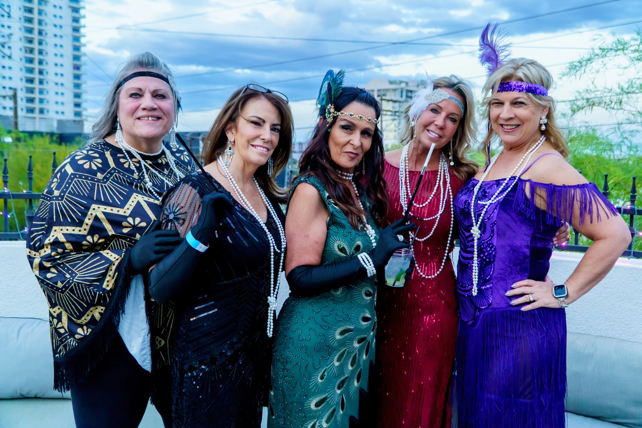 Six women dressed in glamorous, vintage-style outfits with pearl jewelry, posing together outdoors on a rooftop with city buildings in the background, some wearing feathered headbands and boas, during daytime.