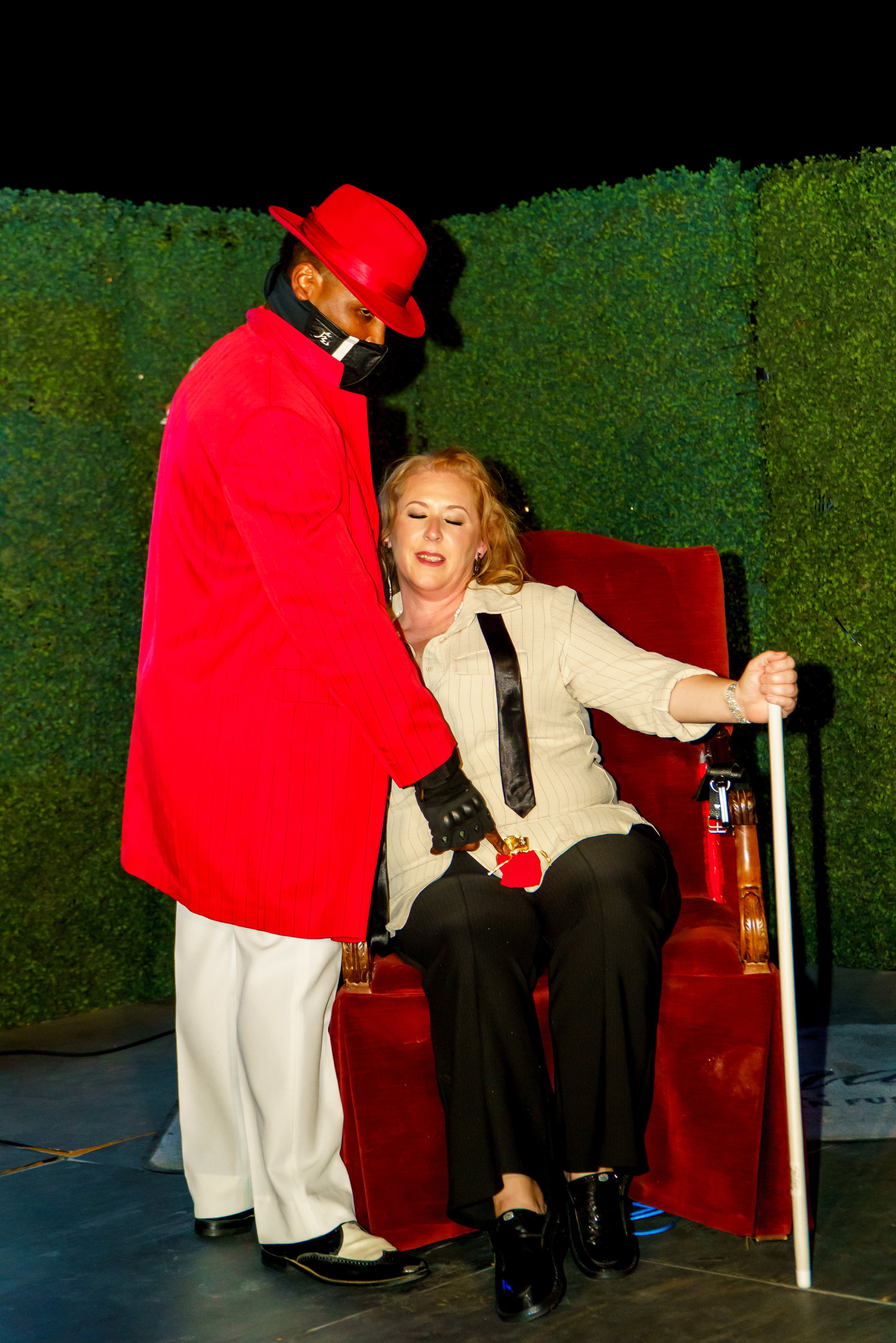 A woman with blonde hair, sitting in a red velvet armchair, is being attended to by a man dressed in a red suit, red hat, white pants, and a black face mask. She has her eyes closed and appears relaxed.