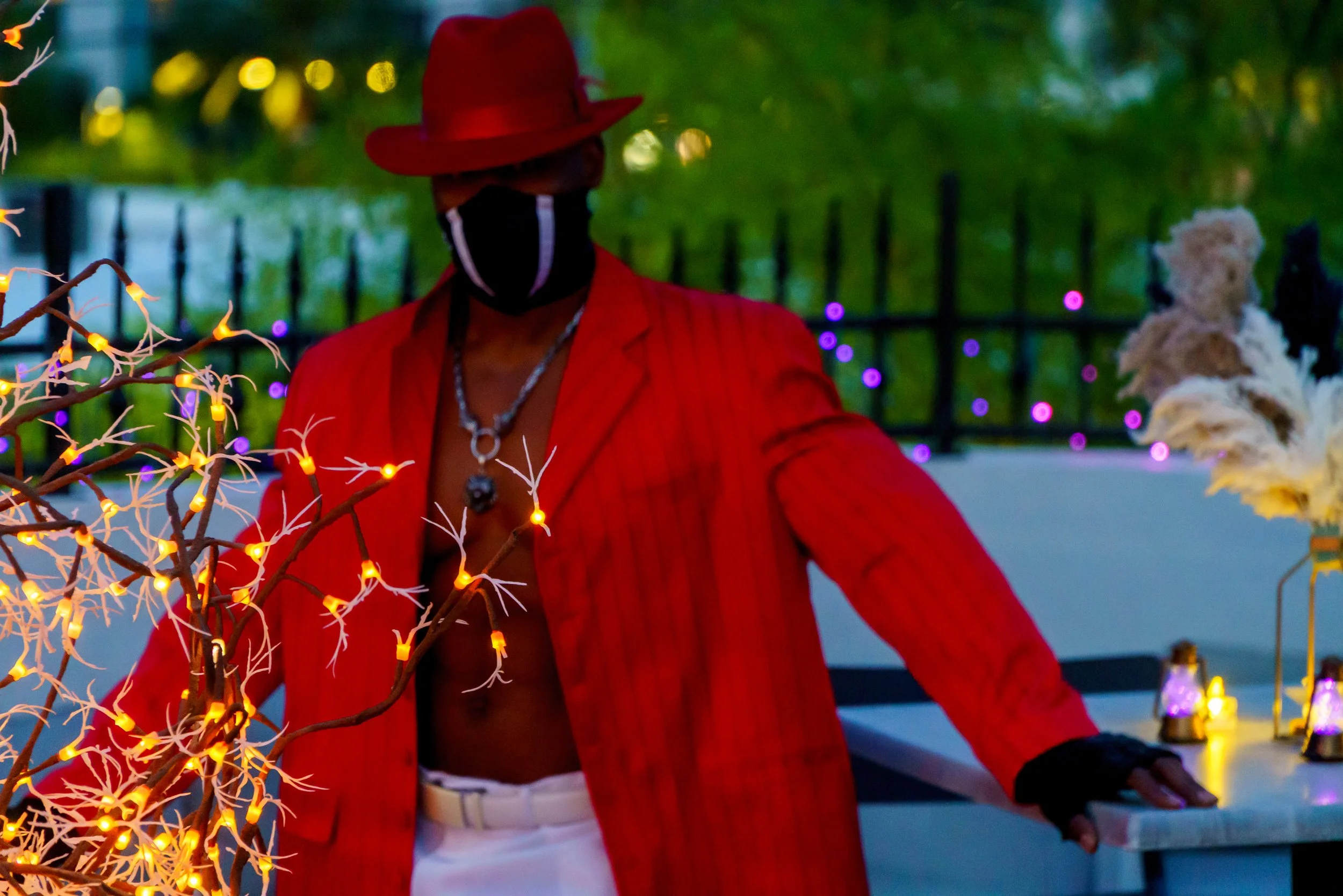 A man wearing a red hat, red jacket, and silver necklace, standing outdoors at dusk near a fence decorated with purple lights, with a small tree with orange lights in the foreground.
