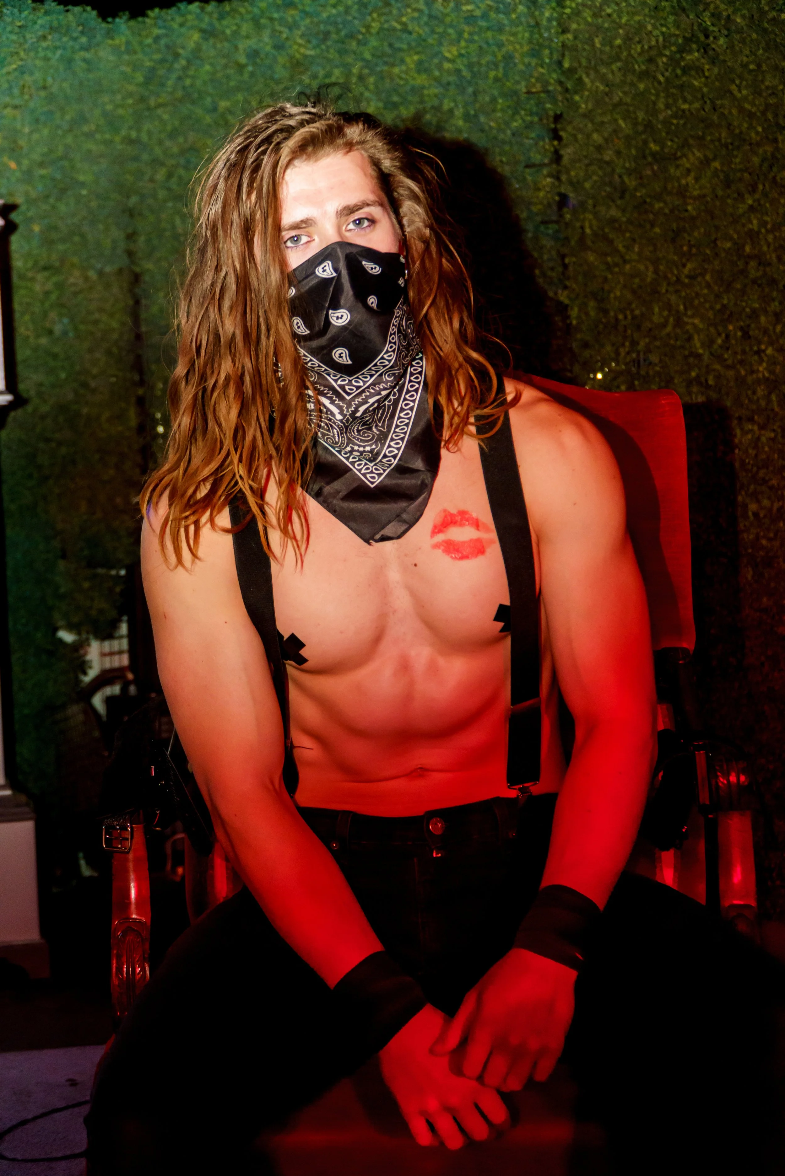 A person with long wavy hair wearing a black bandana mask, black suspenders, gloves, and no shirt, sitting with hands clasped on their lap, with a red lipstick kiss mark on their chest, in a dimly lit setting.