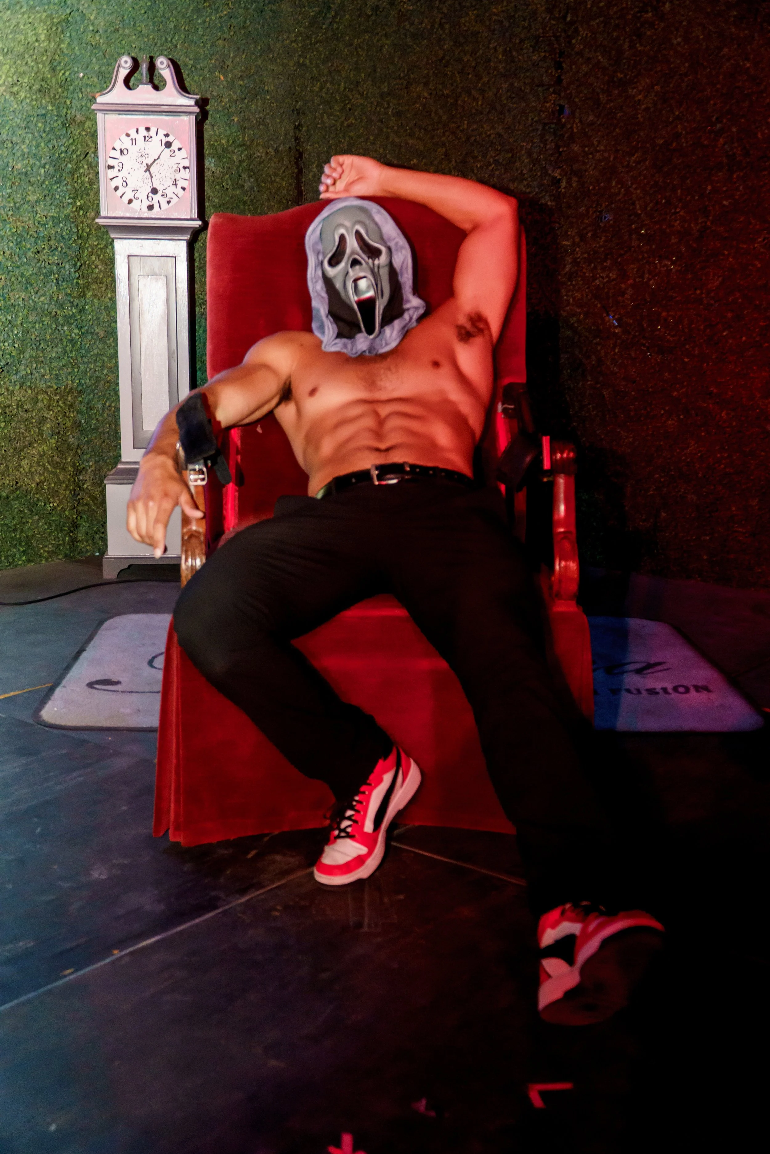 A shirtless man lying on a red lounge chair, wearing a ghost face mask, black pants, and red and white sneakers, with a clock showing 12:24 in the background.