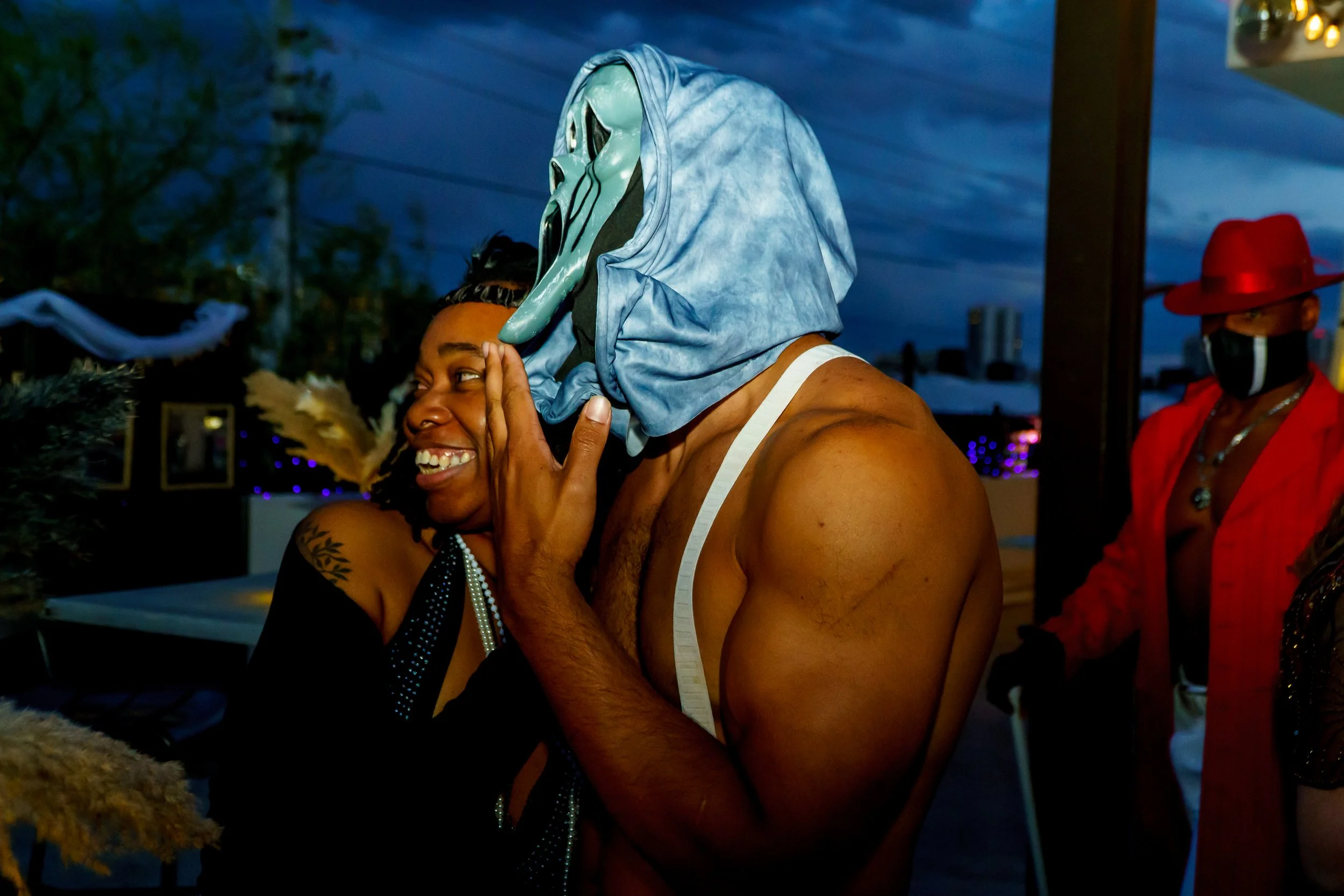 Two people at a party, one wearing a spooky white mask and a blue hood, and the other smiling with hand on face, the scene is outdoors during evening with blue sky.