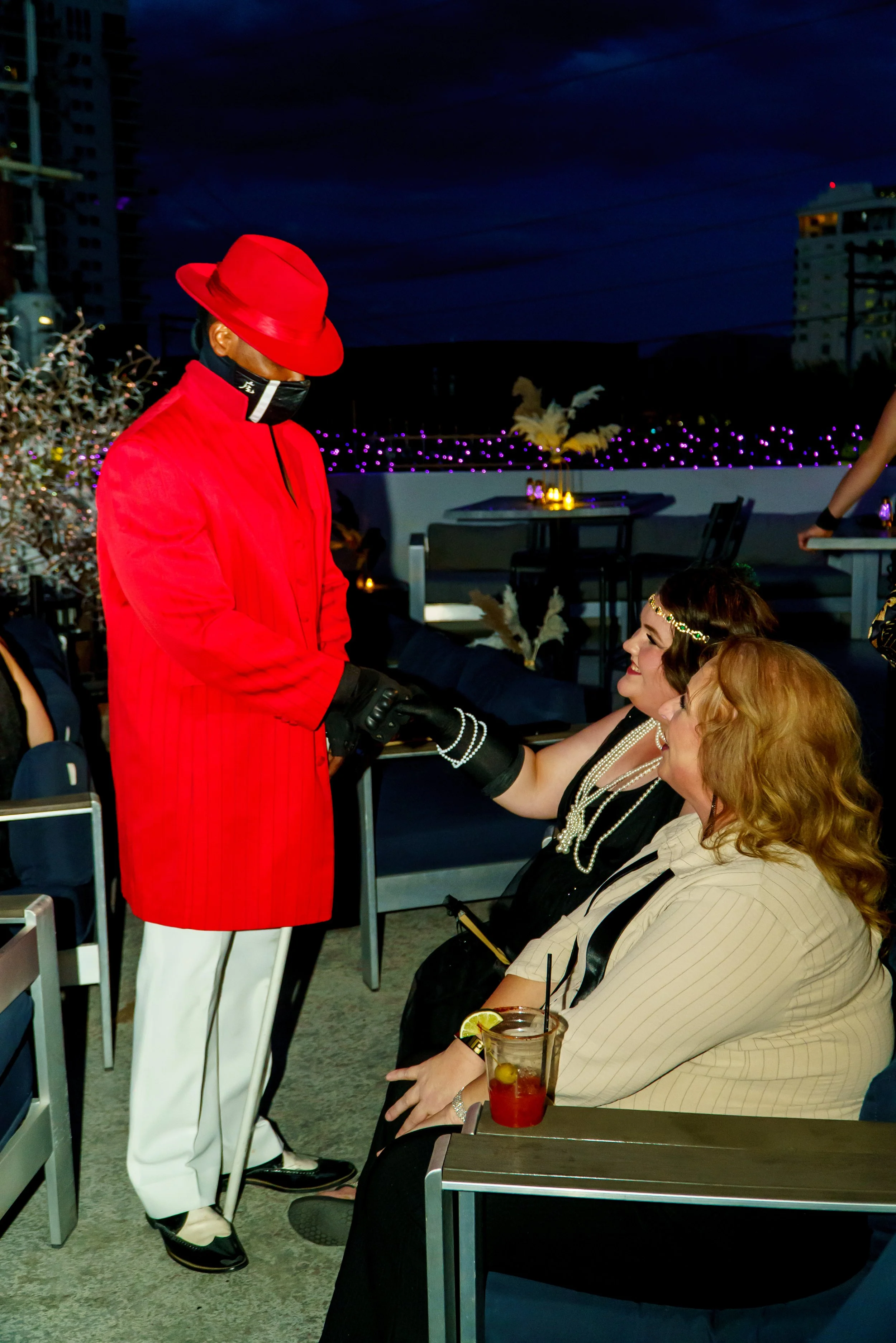 A person wearing a red hat, red coat, and a face mask standing and holding hands with a woman sitting at a table. The woman has curly hair and is wearing a black dress with pearl accessories. The scene is outdoors at night with city buildings and pur
