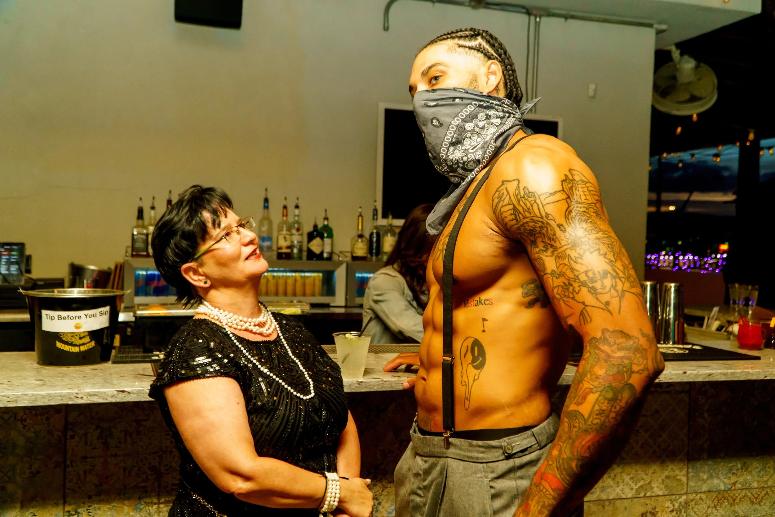 A woman in a black sequin dress and pearl jewelry is smiling and looking up at a shirtless man with tattoos and a black bandana covering his face, standing at a bar in a dimly lit setting with liquor bottles in the background.