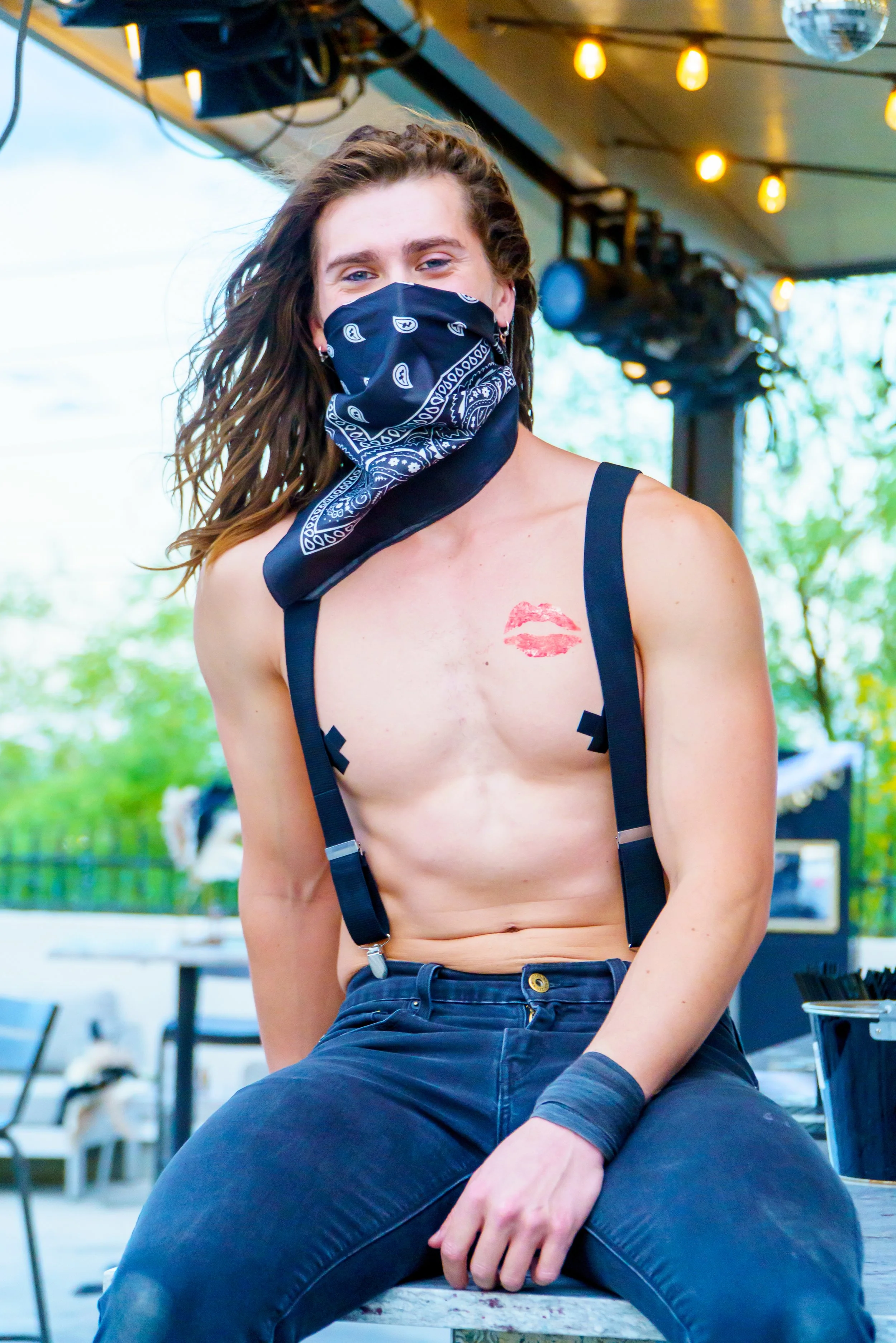 A person with long hair wearing a black bandana mask, a lipstick mark on chest, black suspenders, and dark pants, sitting outdoors with string lights overhead and blurred background of trees and furniture.