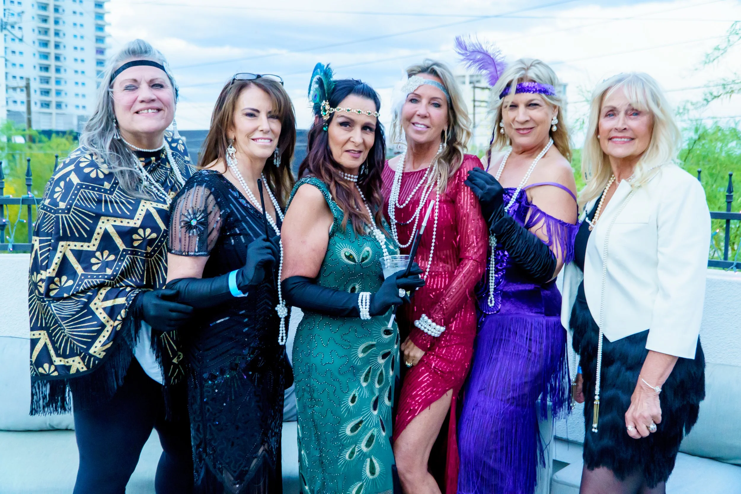 Group of six women dressed in vintage 1920s flapper costumes, accessorized with pearls, headbands, feathers, and gloves, standing outdoors on a balcony with city buildings in the background.