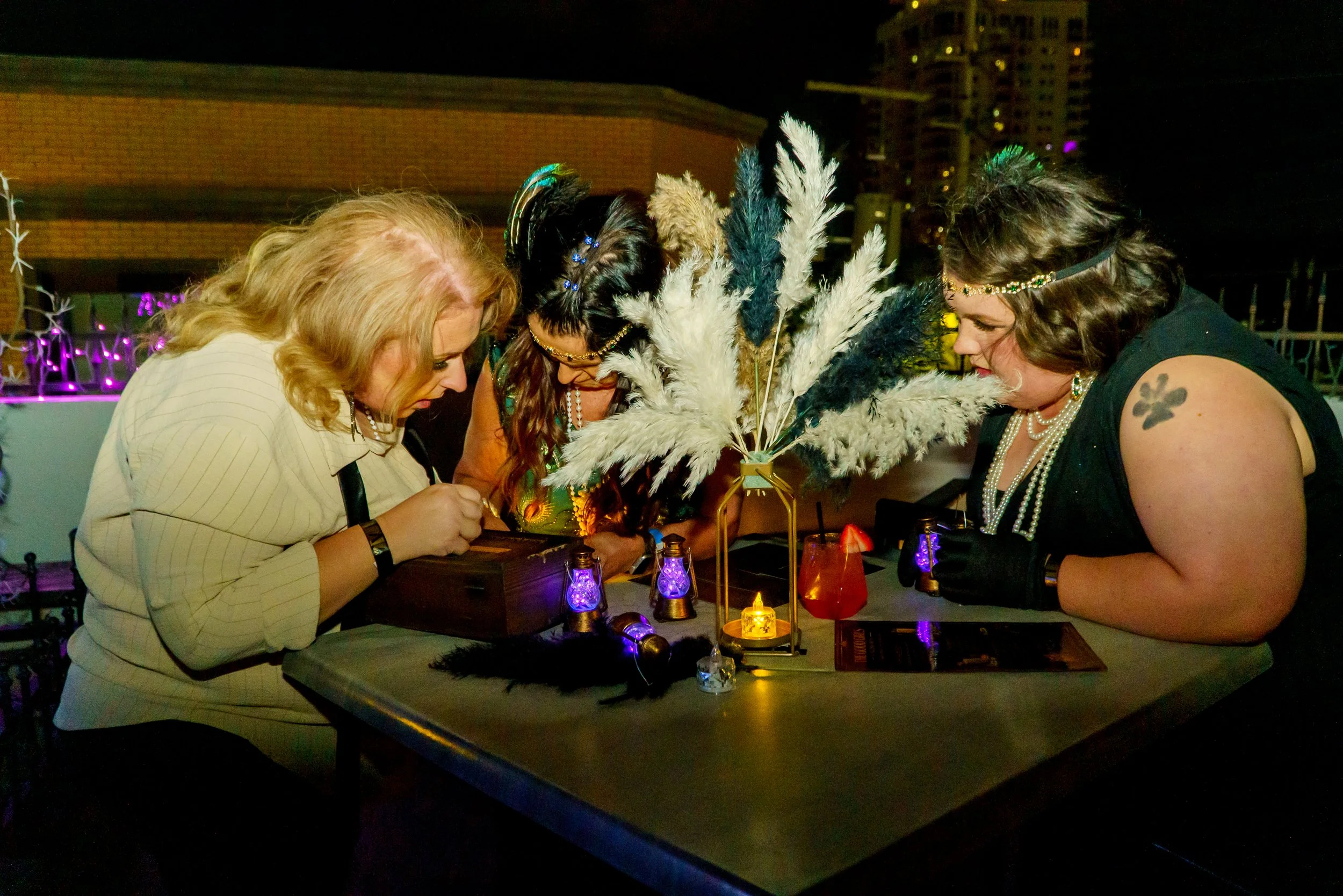 Three women are gathered around a table at night, examining or signing something with a festive atmosphere, decorative feathers, candles, and colorful lanterns illuminating the scene.