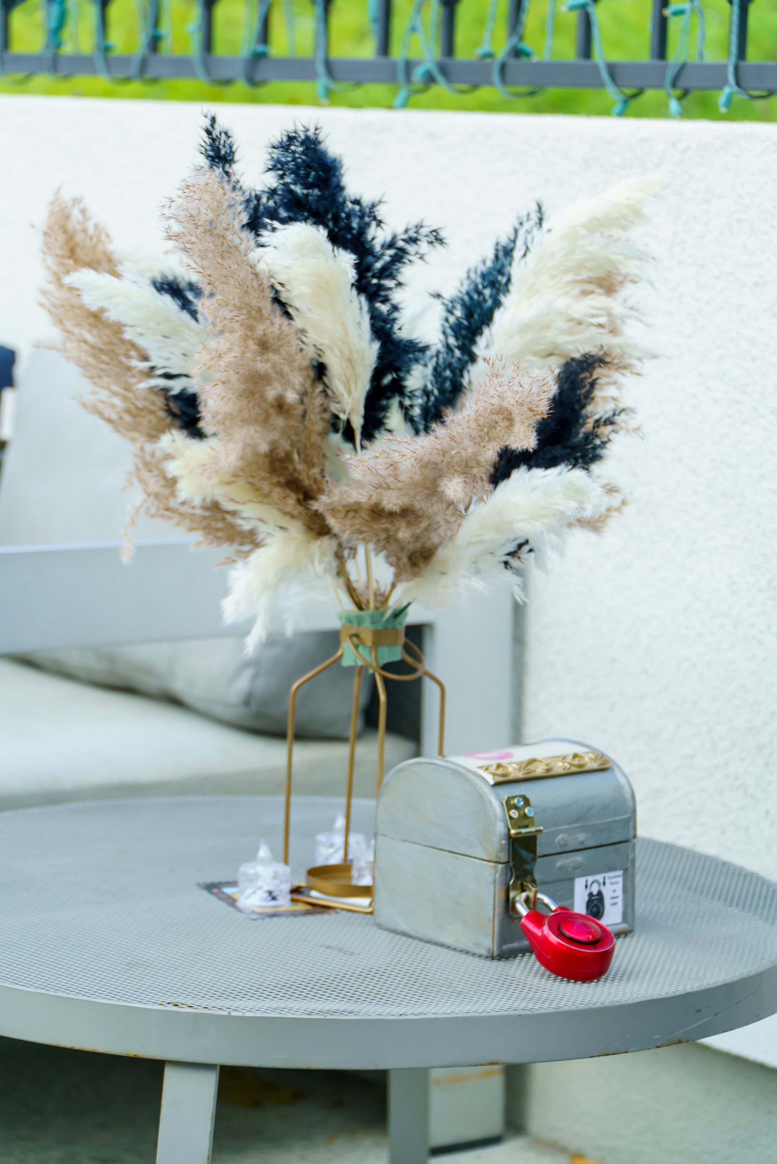 A display of feathers arranged on a circular table, with a metallic gift box, a red object, and some papers or cards also on the table. The feathers are in shades of white, beige, and black. The background includes a textured white wall and a railing