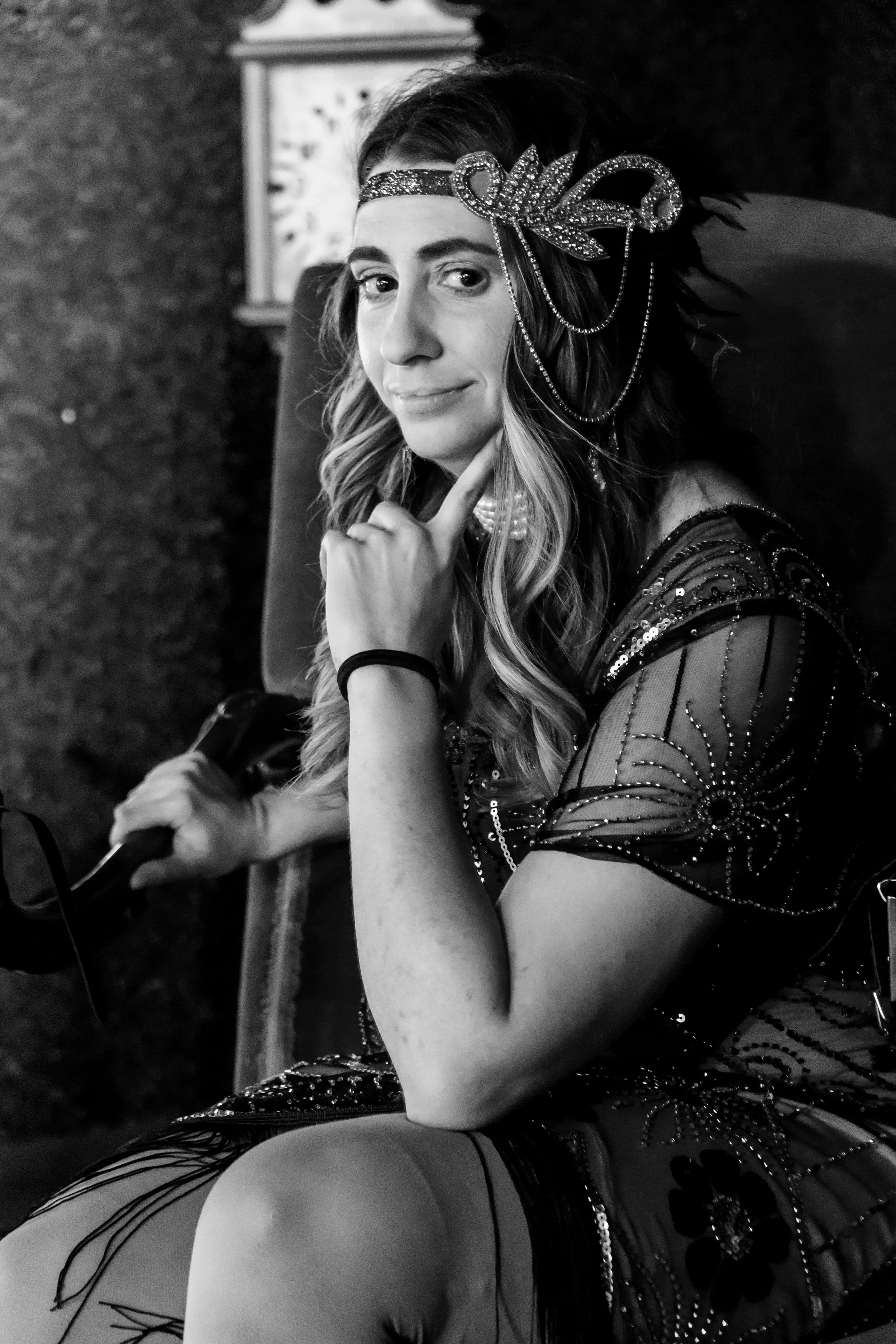 A woman dressed in a vintage, flapper-style costume, sitting on a chair with her chin resting on her hand, wearing a bejeweled headband and a dress with intricate beading and details, black and white photo.
