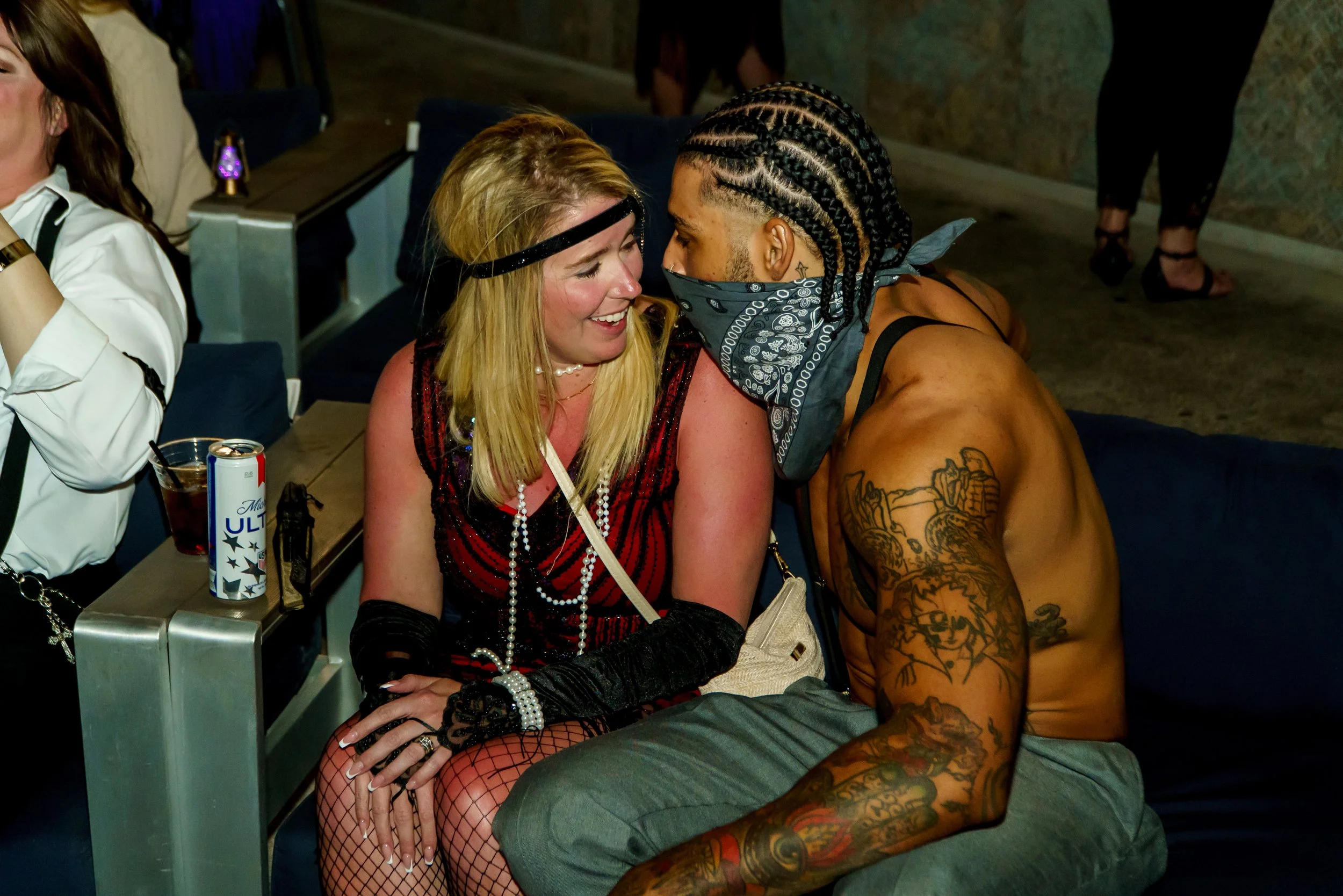 A couple sitting close together, with their foreheads touching, smiling at each other. The woman is dressed in a red and black outfit with fishnet stockings and long black gloves, wearing a headband and pearl necklace. The man is shirtless, wearing a