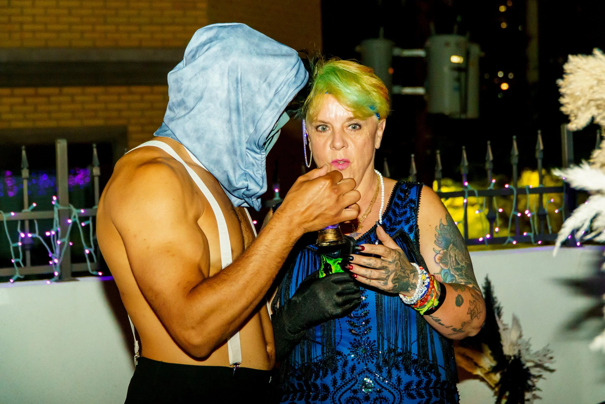 A woman with short, multicolored hair wearing a blue dress and jewelry, holding a wine glass, and standing next to a shirtless person with a hood over their head at night.