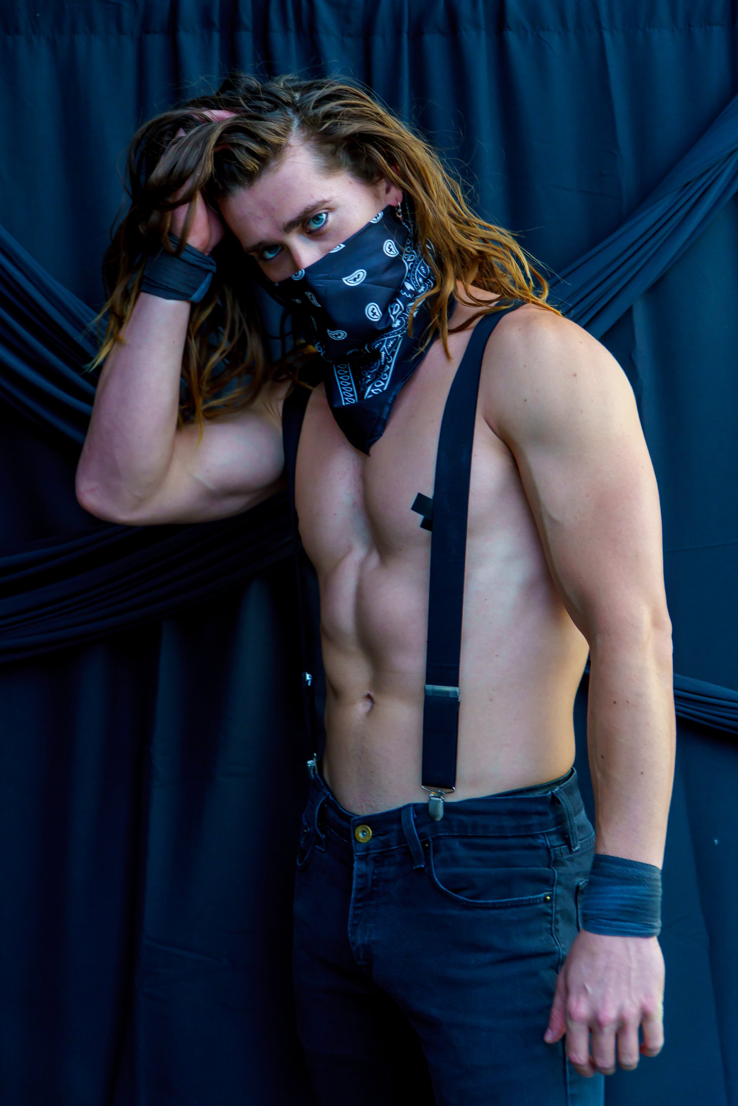 A woman with curly hair, wearing a black bandana over her nose and mouth, a black strap top, black jeans, and dark socks, stands in front of a black backdrop with draped fabric. She has her right hand in her hair and is looking directly at the camera