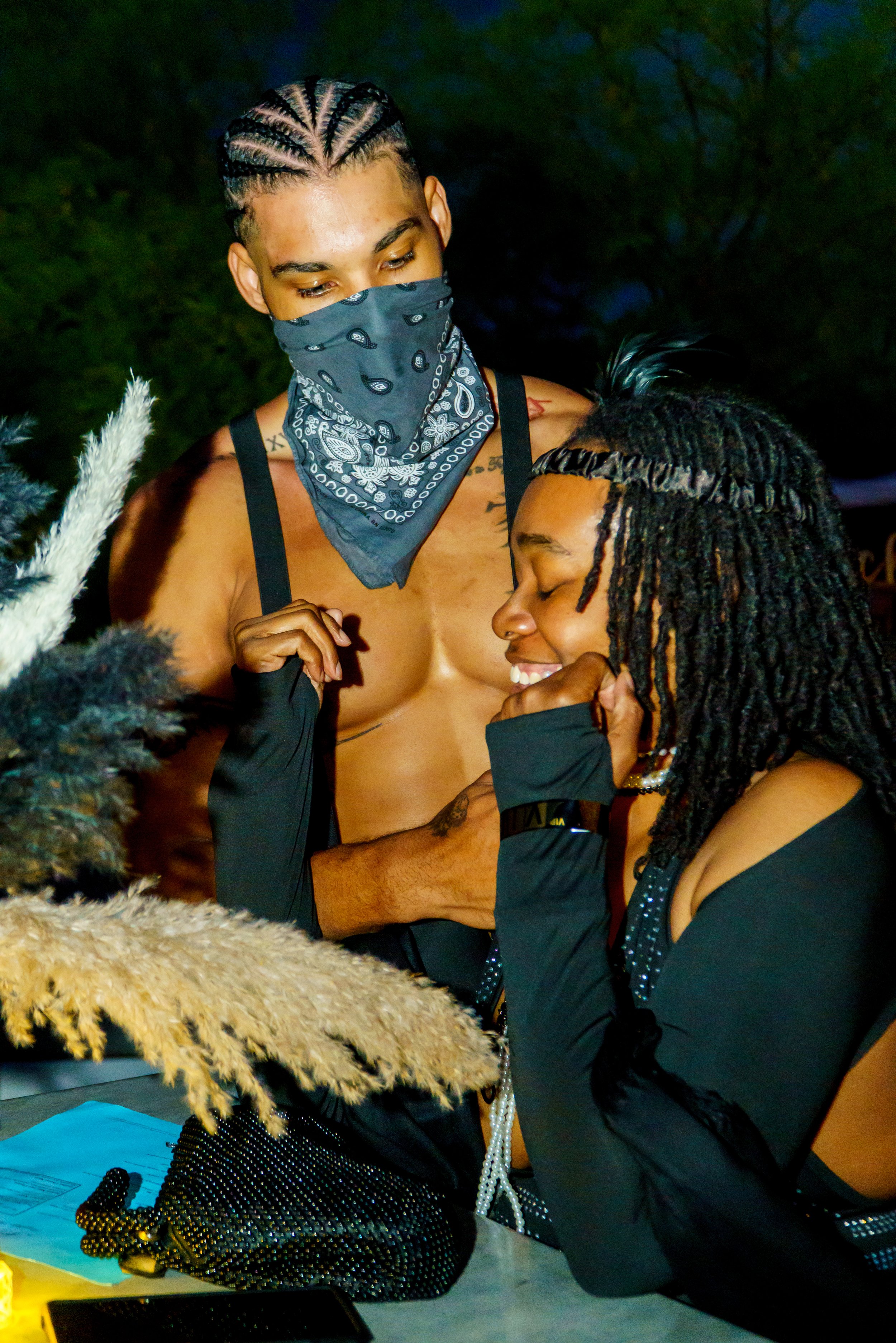 Two women, one with a braid and the other with dreadlocks, are embracing and smiling in a close, intimate moment. One woman is wearing a bandana over her face, and they are surrounded by various objects including a black beaded purse, dried pampas gr