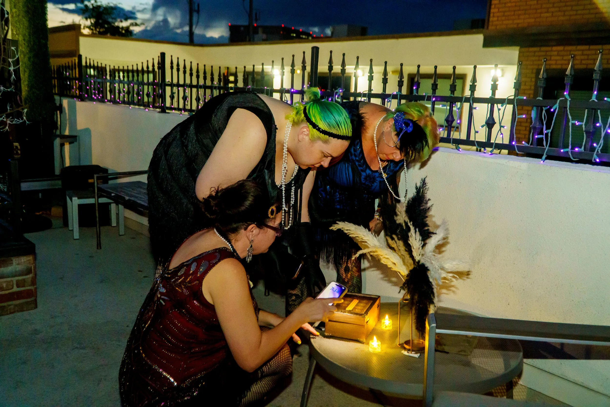 Three women dressed in colorful, glamorous outfits with accessories such as pearl necklaces and headbands, are gathered around a small table with candles, on an outdoor balcony or patio at night, engaged in conversation or an activity.
