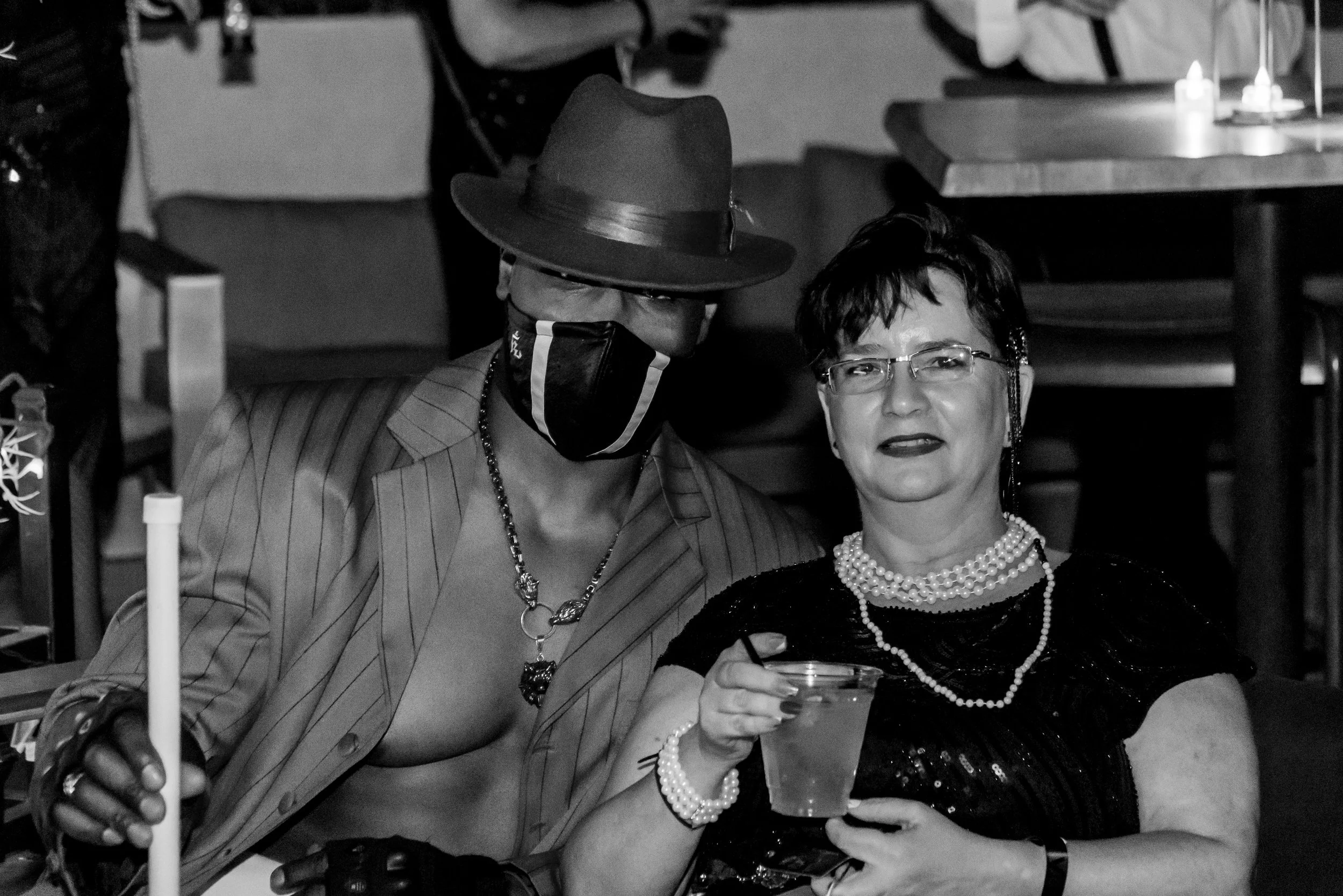 Two women seated at a social gathering, one wearing a striped blazer, a large hat, a face mask, and jewelry, and the other wearing glasses and a pearl necklace, holding a drink.