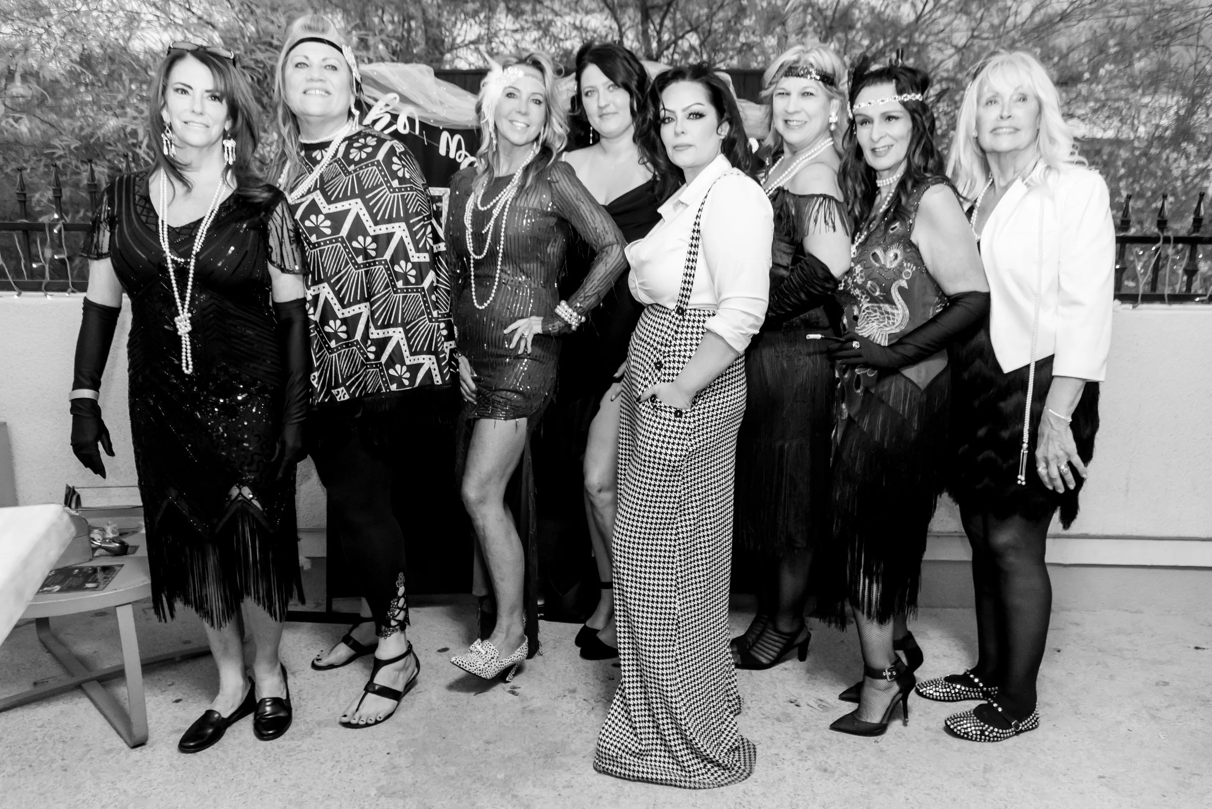A group of nine women dressed in vintage 1920s style outfits, standing together outdoors near a fence with trees in the background. They are wearing flapper-style dresses, headbands, long necklaces, and gloves, and are posed for a photograph.
