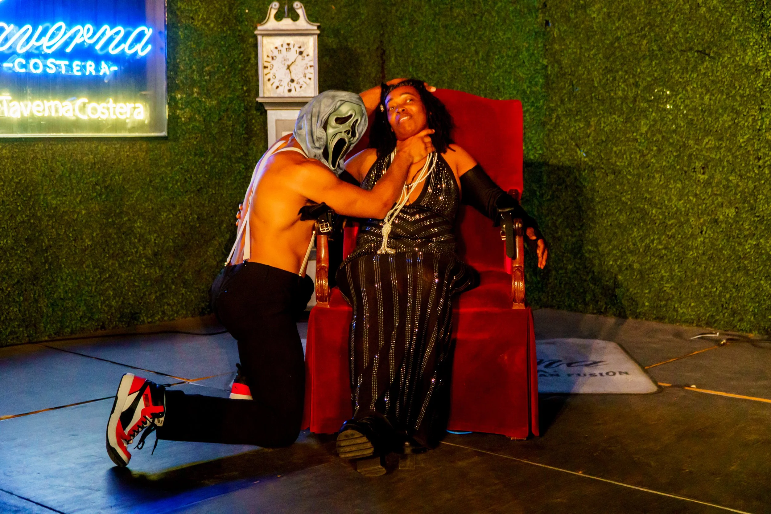 A person dressed in a Halloween costume with a ghost face mask kneels next to a woman sitting on a red chair. The woman, dressed in a black outfit with jewelry, poses with a playful expression, resting her head on her hand. There is a wall with green