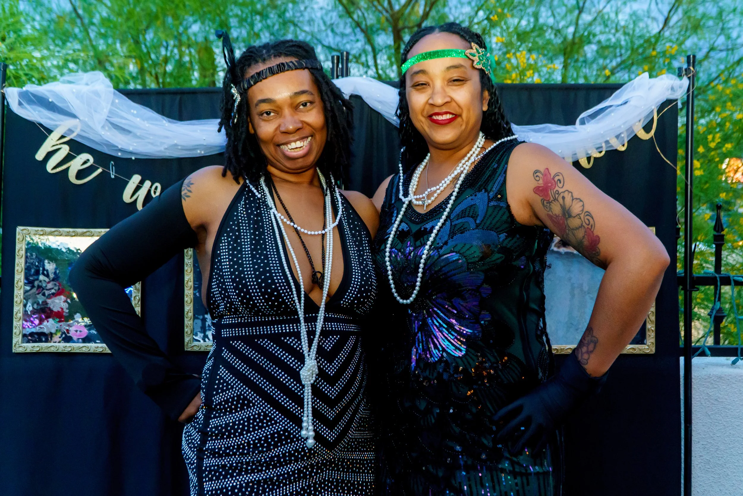 Two women smiling and dressed in black, wearing pearl necklaces, posing at an outdoor event with a backdrop of trees and a black banner with white and gold decorations.