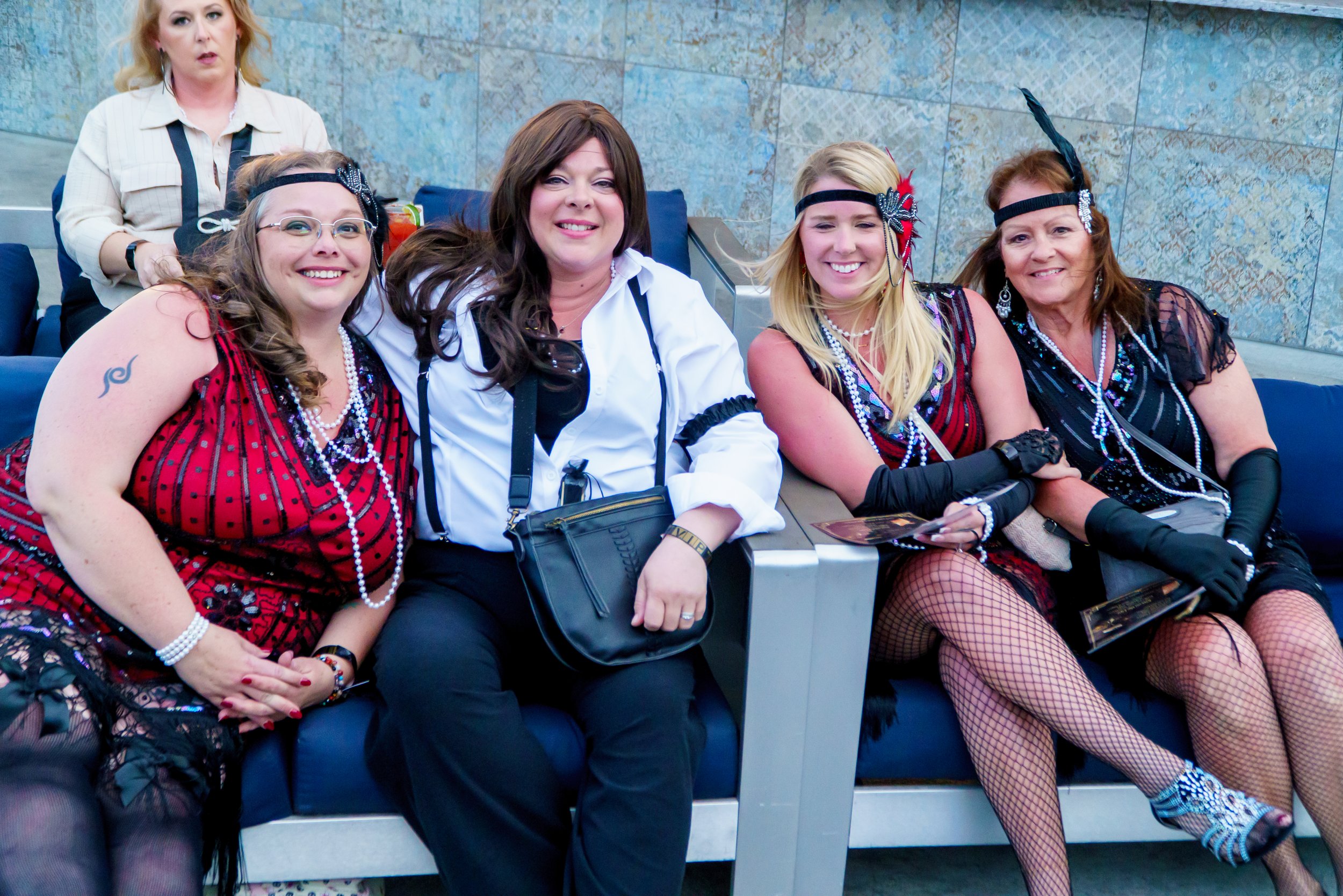 Group of five women sitting on blue seats in an indoor venue, dressed in colorful costumes with accessories, some smiling and others with neutral expressions, against a stone wall background.