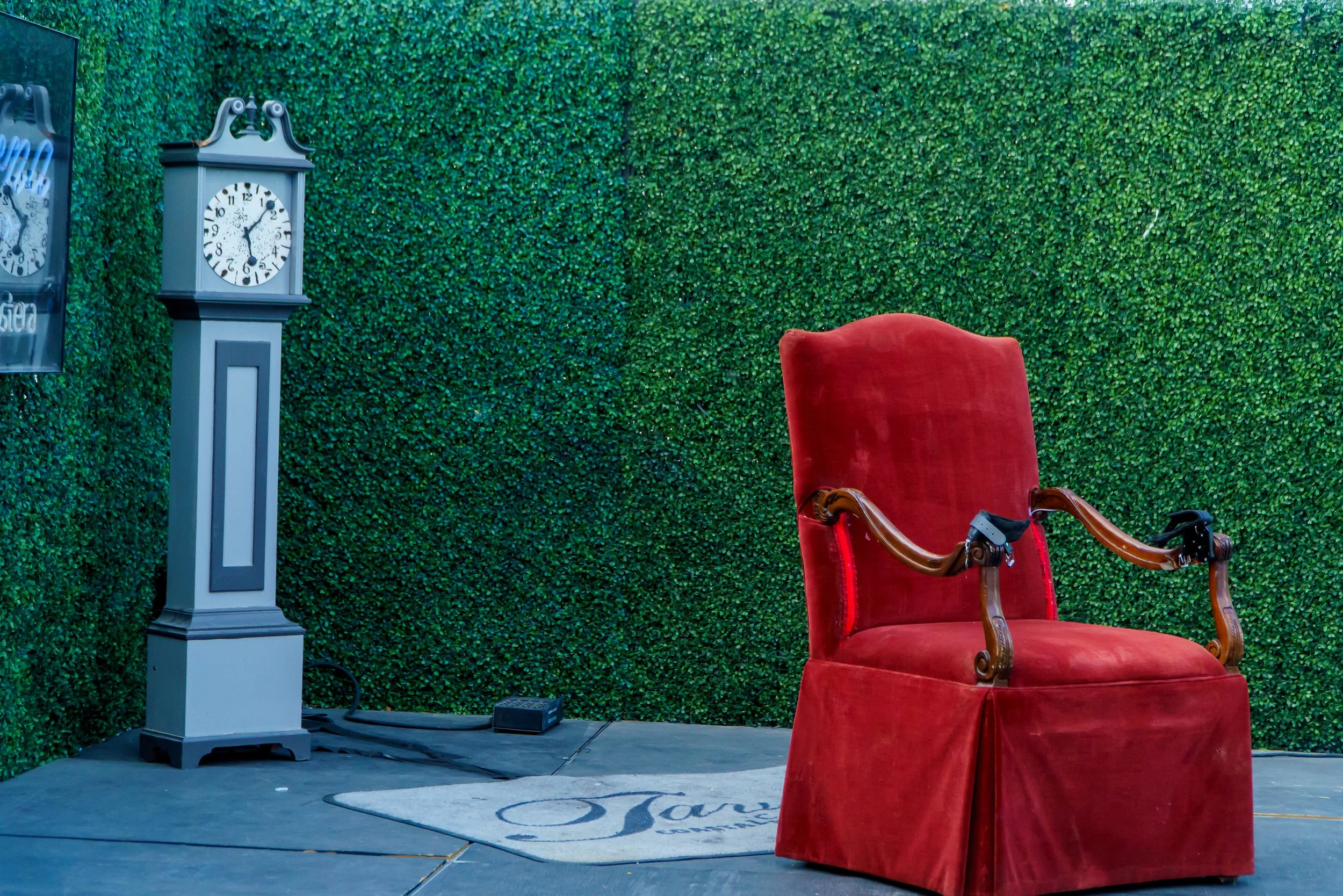 A red velvet armchair with wooden armrests placed on a stage with a decorated backdrop of green foliage. To the left, there is a gray antique-style clock and a small electronic device on the floor.