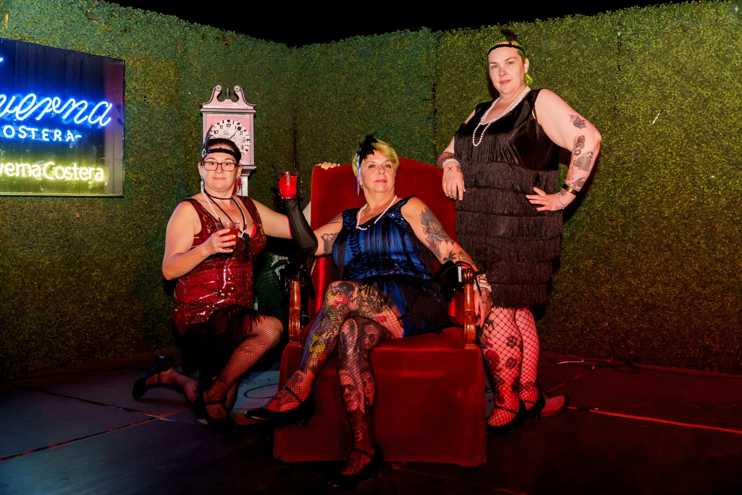 Three women in vintage-style clothing and fishnet stockings, with tattoos, in a dimly lit room with green hedge wall and neon sign, seated and standing around a red velvet chair, holding drinks.