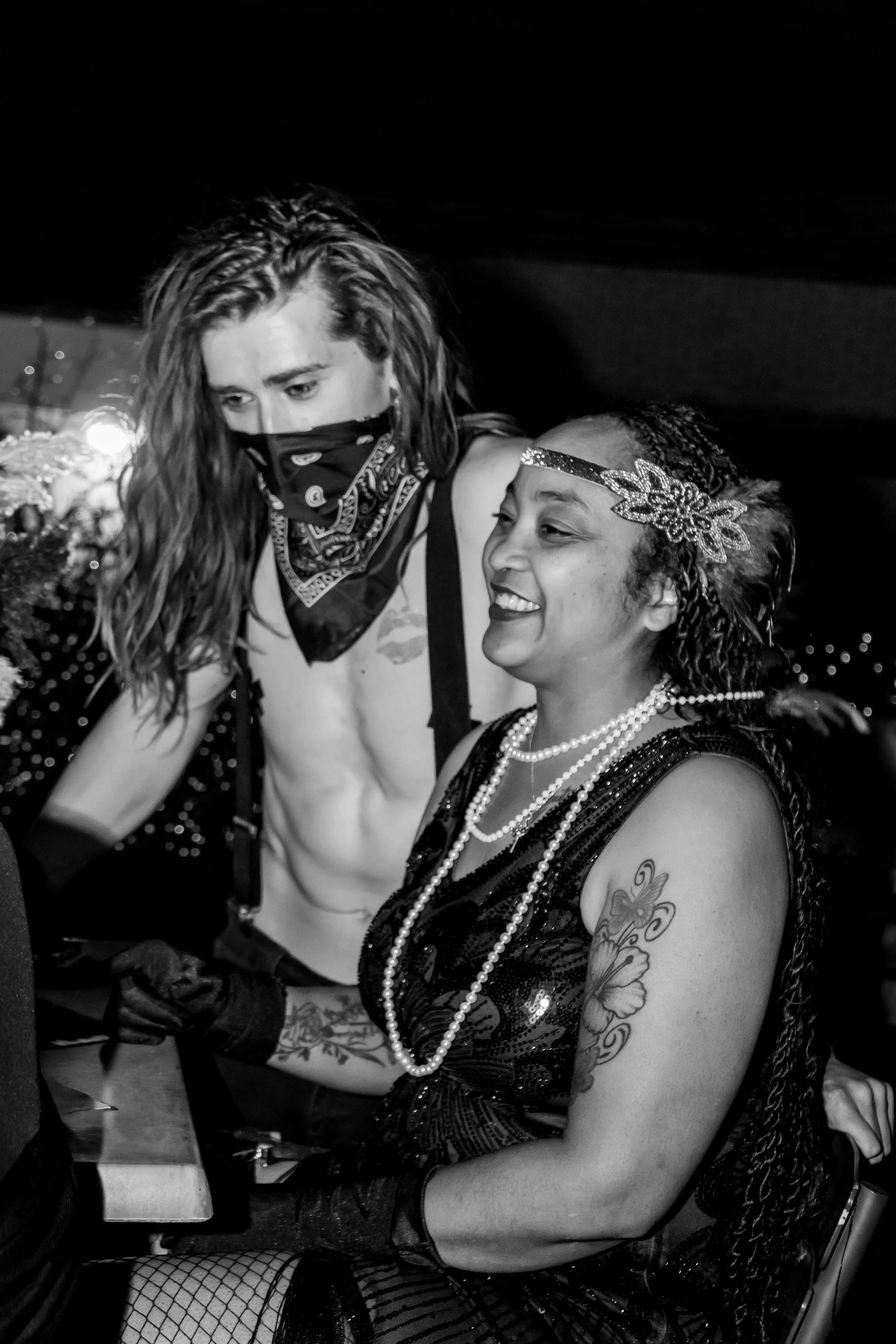 Two women dressed in costume, one wearing a floral headband, pearl necklace, and tattoos on her arm, the other wearing a bandana over her face, with fishnet stockings visible, in a dark setting.