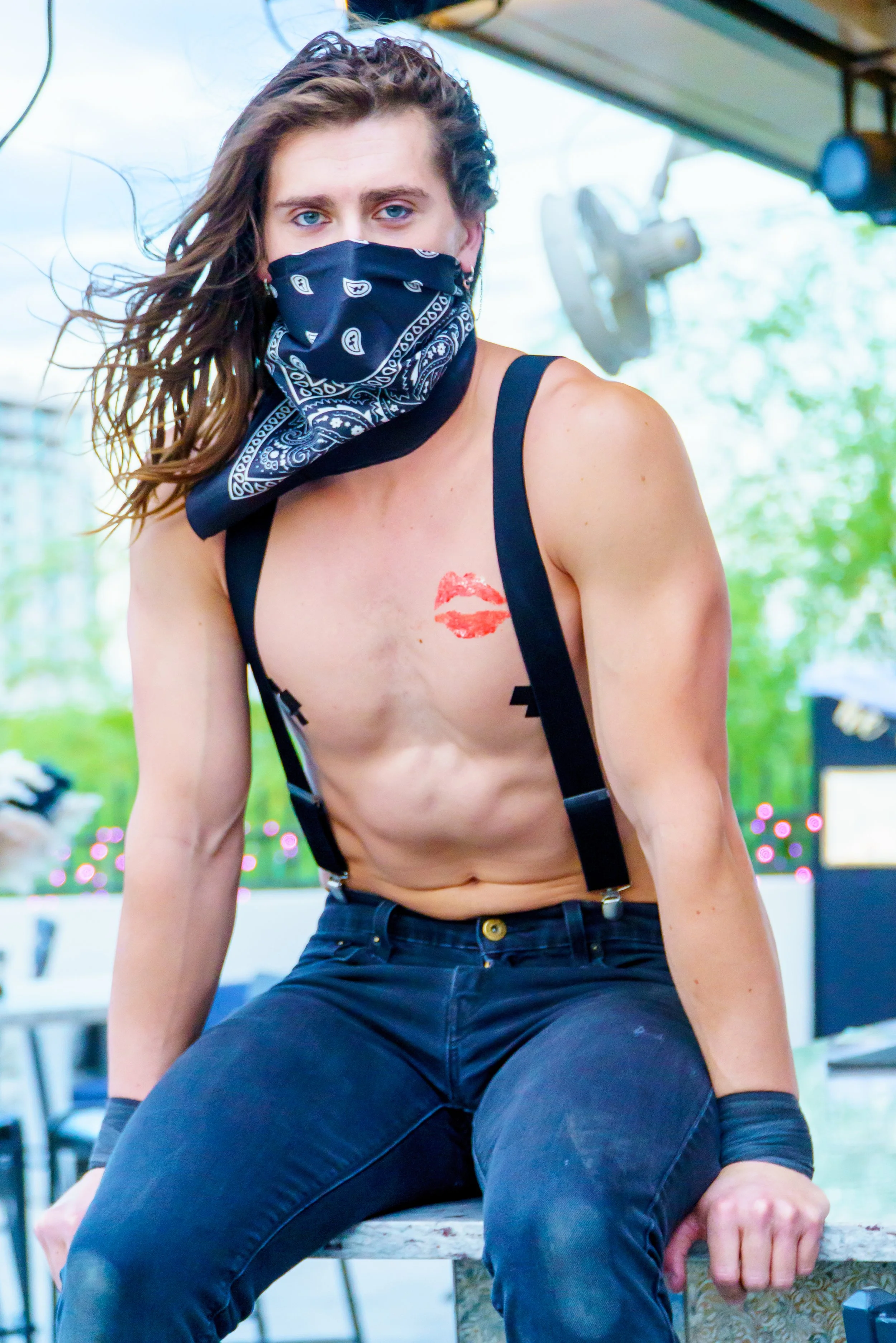 Person with long hair wearing a black bandana mask, exposed upper body with a lipstick kiss mark on chest, in a room with large windows, greenery outside, and studio equipment in the background.