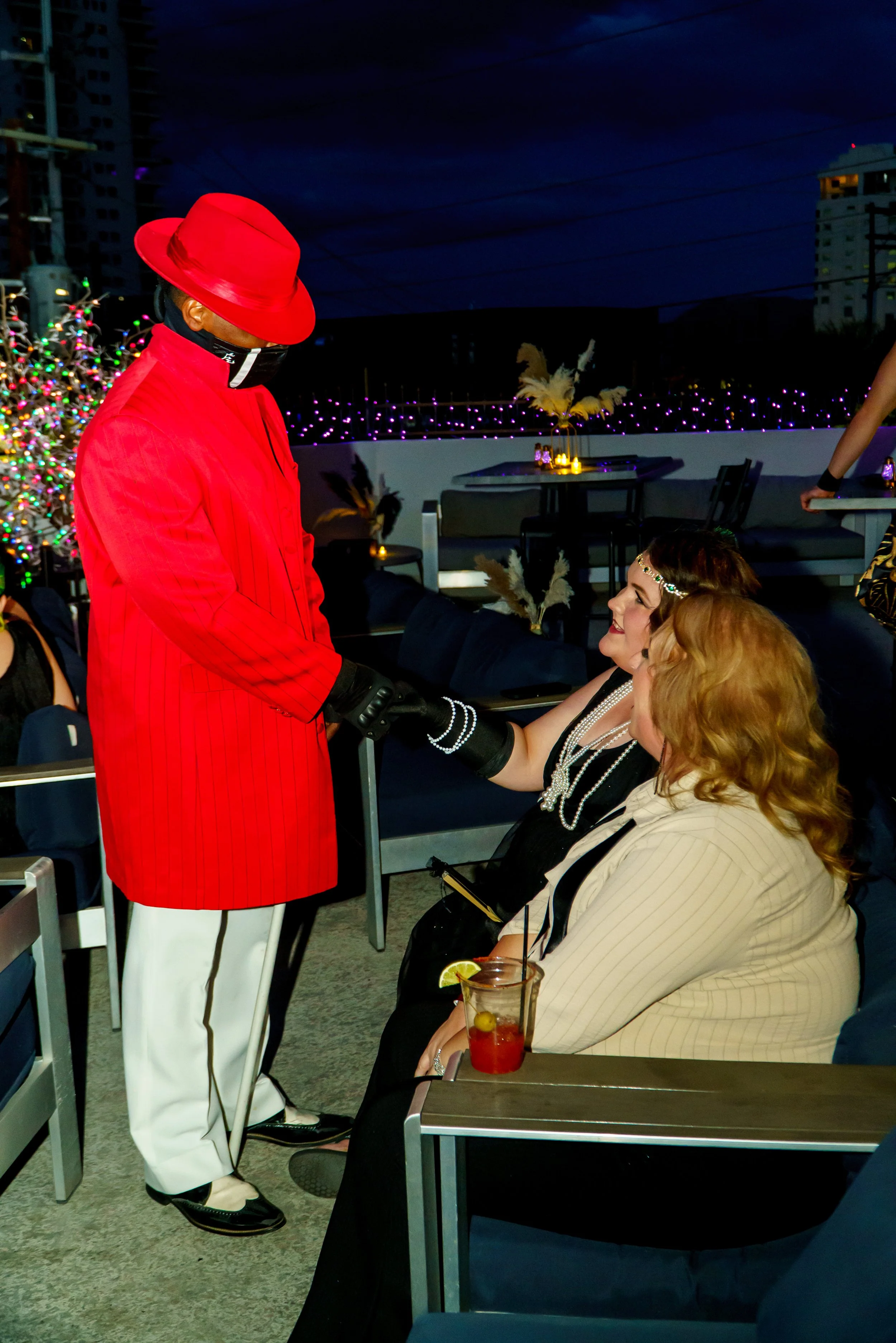 Man dressed in red coat, white pants, red hat, and black gloves shaking hands with woman in black dress with pearl necklace at outdoor evening event, decorated with colorful Christmas lights and feathers.