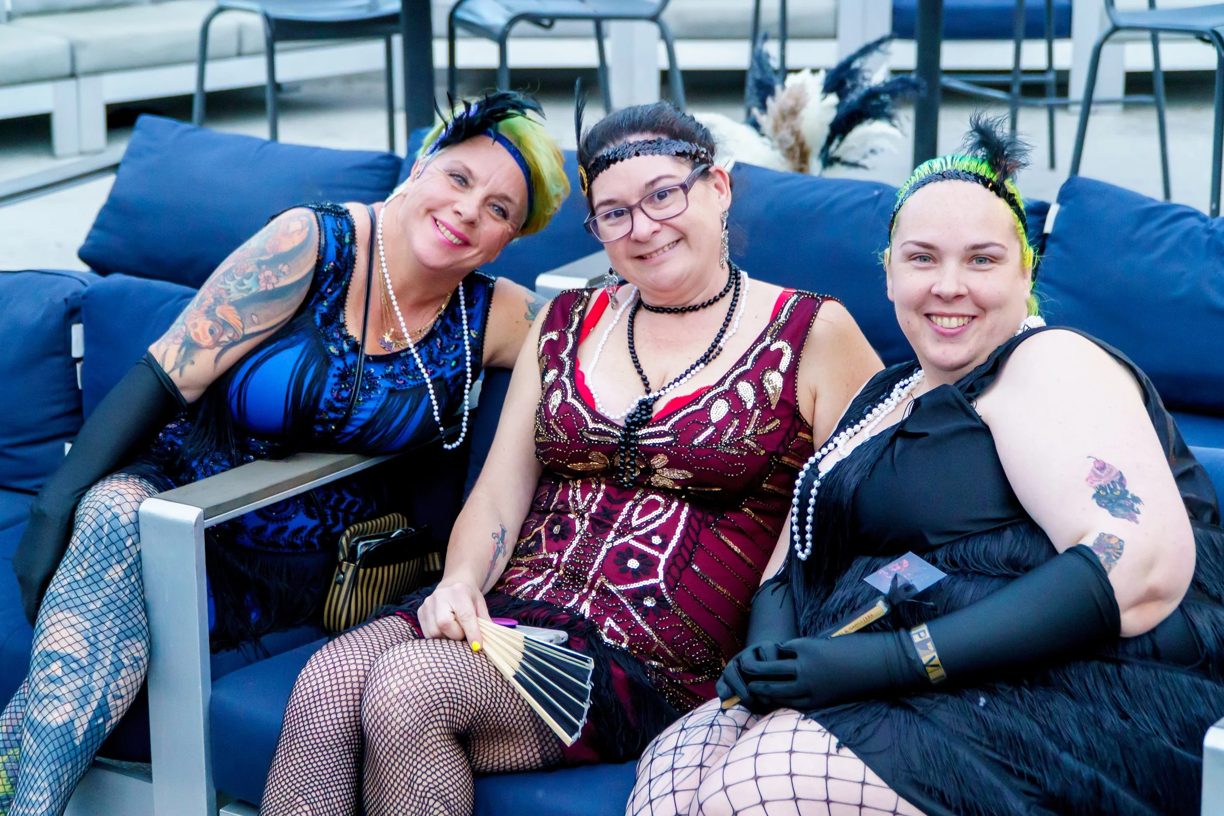 Three women dressed in burlesque or carnival costumes sitting on a blue outdoor couch, smiling at the camera.